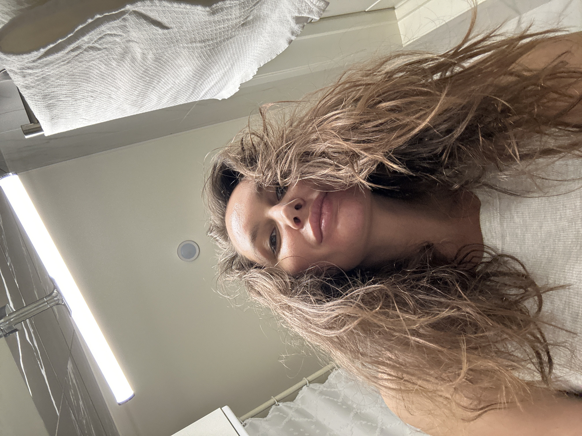 An overhead, low-angle shot captures a woman looking directly downwards towards the camera, likely in a bathroom. Her face is visible in the center, framed by her voluminous, light brown, wavy hair, which appears wet or disheveled and spreads across the frame. Her lips are slightly parted, and her gaze is neutral.
The background consists of light-colored walls and ceiling. In the upper left, a white, textured towel hangs on a silver-toned metal fixture. A bright, vertical rectangular LED light bar is prominent on the left, casting a strong illumination onto a dark, possibly tiled, wall surface. A small, circular recessed light fixture is also visible on the ceiling in the upper right. A white, textured pattern, possibly from a shower curtain or a textured wall, can be seen along the right and bottom edges of the frame, partially covered by the woman's hair. The lighting is artificial and bright. There is no visible text. The scene is consistent with an indoor setting, specifically a bathroom, in Moscow, Russia.