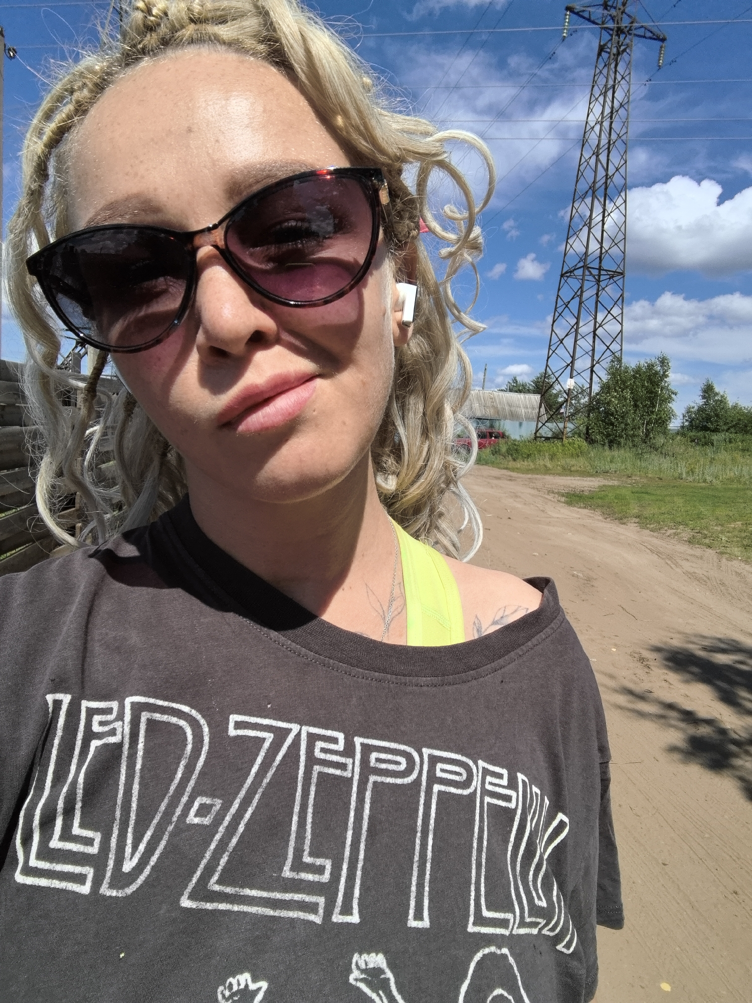 The media file shows a close-up, selfie-style shot of a woman with blonde, braided hair, wearing sunglasses and a dark t-shirt with a "LED-ZEPPELIN" logo. She is outdoors, with a power line tower and a building visible in the background. The scene appears to be a rural setting in Noviy, Russia, with a dirt road and vegetation. The weather seems to be sunny with a blue sky and some clouds. The woman is looking at the camera with a neutral expression. The time of day is likely afternoon, given the shadows. The image captures a casual, everyday moment, possibly during an outdoor activity.