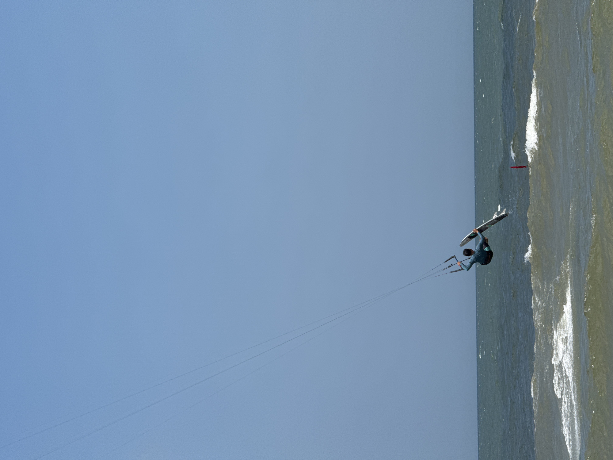 An individual kite surfer is captured mid-air over the ocean at Mũi Né, Vietnam. The surfer, dressed in a blue wetsuit and green shorts, is positioned on a board and appears to be performing a trick. They are attached to a large kite by lines that extend high into the clear blue sky. The ocean below is choppy with white waves. A single red buoy is visible in the water to the right of the surfer.
