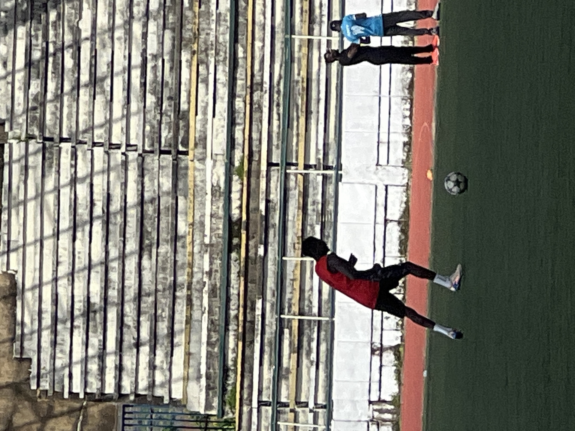 In New Stadium, Nigeria, a soccer player in a red shirt and black shorts is captured mid-stride on a green artificial turf field, with a soccer ball at his feet. He is facing away from the camera, his right leg extended forward. In the background, three other individuals are visible. One, in a black shirt and pants, stands with his arms crossed. Another, in a blue shirt and dark pants, is looking towards the player. Behind them, the weathered concrete bleachers of a stadium rise up, some sections showing signs of disrepair with green overgrowth. The scene suggests a training or practice session in an outdoor sporting venue.