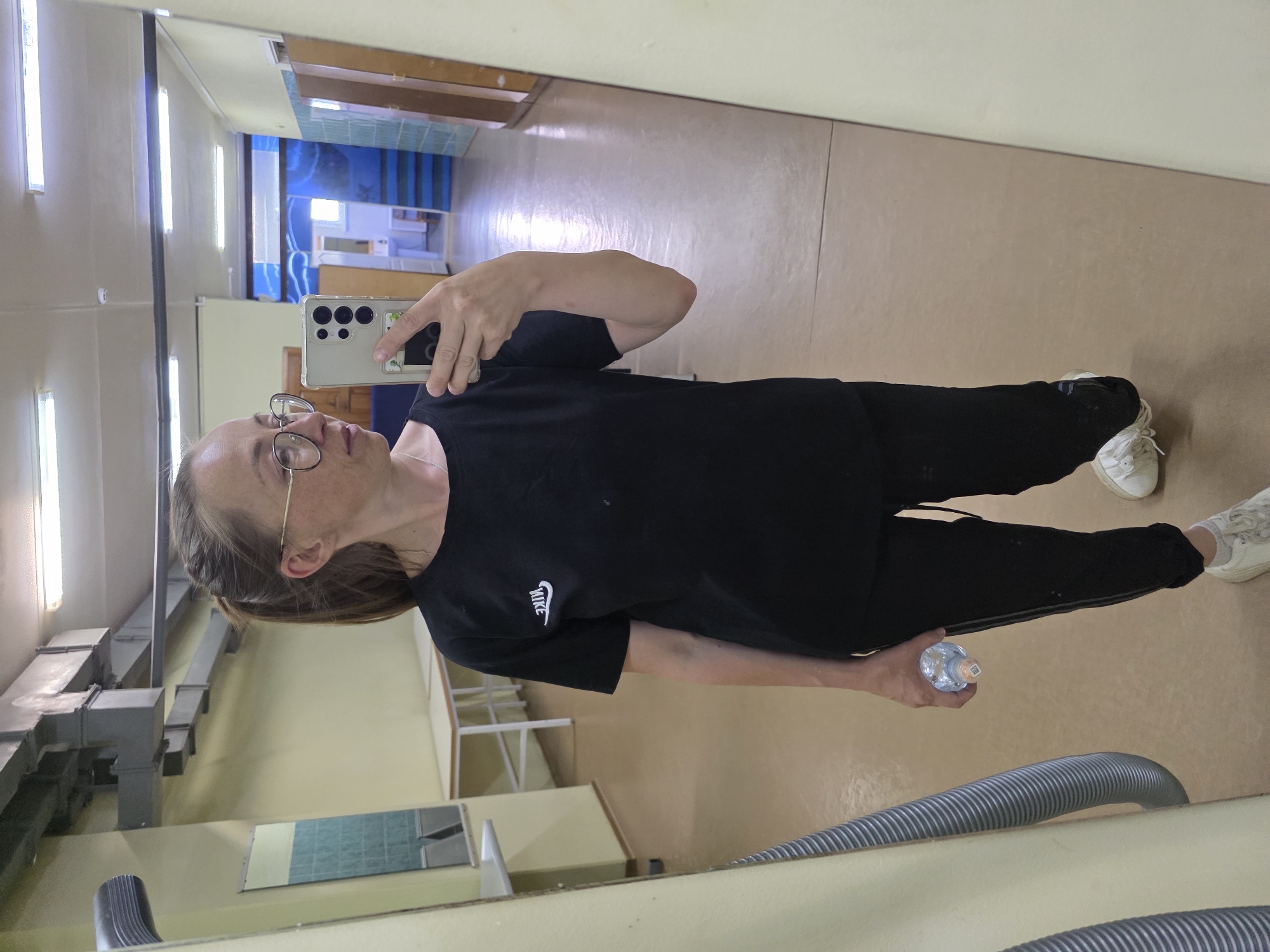 A woman is taking a selfie in a mirror. She is wearing a black T-shirt with a white Nike logo, black pants, and white sneakers. She is holding a water bottle in her left hand and a smartphone in her right. She has glasses and her hair is pulled back.
The reflection in the mirror shows a hallway with fluorescent lights on the ceiling. There is a blue wall with windows at the end of the hallway. A heating duct runs along the side of the hallway. The floor is a beige color.
Based on the available information, it is not possible to confirm the location in the city of Chaikovsky, Russia.