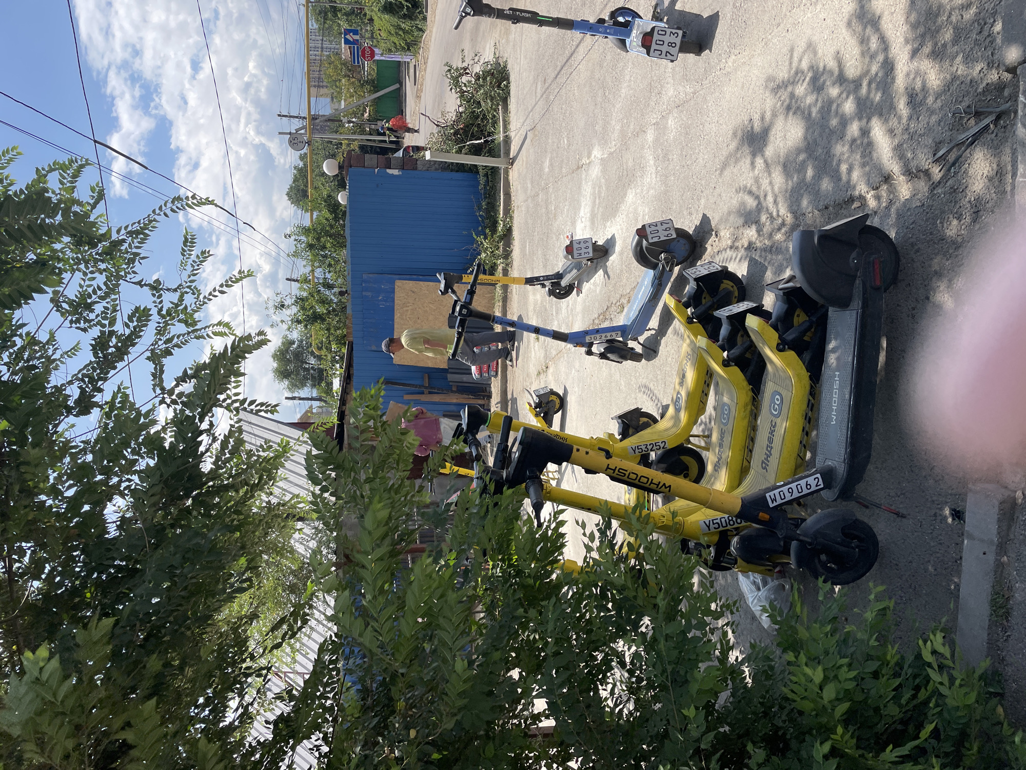A street scene in Almaty, Kazakhstan shows a row of yellow and blue electric scooters parked on a paved surface under a blue sky with scattered clouds. Several of the yellow scooters are branded with "WHOOSH" and "Яндекс.GO." One blue scooter is branded "JET FUSH." The scooters are lined up in front of a blue corrugated metal fence. A man in a light-colored shirt and dark pants is walking away from the scooters towards the fence. Another person in a pink shirt and dark pants stands behind him, partially obscured by a tree. A third person in a yellow shirt is visible walking away from the camera in the background. Green foliage from a nearby tree casts shadows across the scene. A stop sign is visible in the distance on the right side of the image.