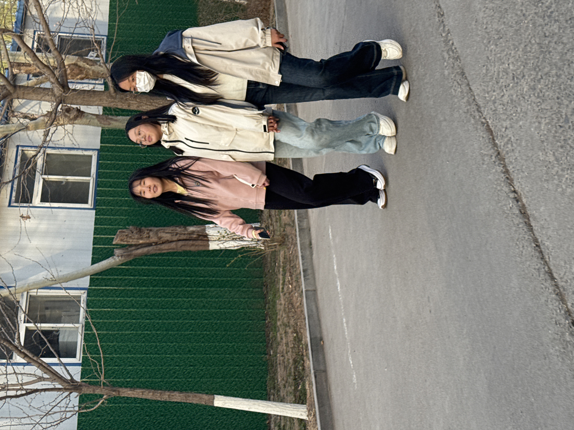 Three young women are walking along a gray asphalt road next to a green and white wall with windows. The wall is next to a tree with bare branches.
The woman closest to the wall is wearing a pink jacket and black pants. The woman in the middle is wearing a white jacket and light blue jeans. The woman farthest from the wall is wearing a beige jacket and dark blue jeans and a white face mask.
The location is Zhongtun, China.