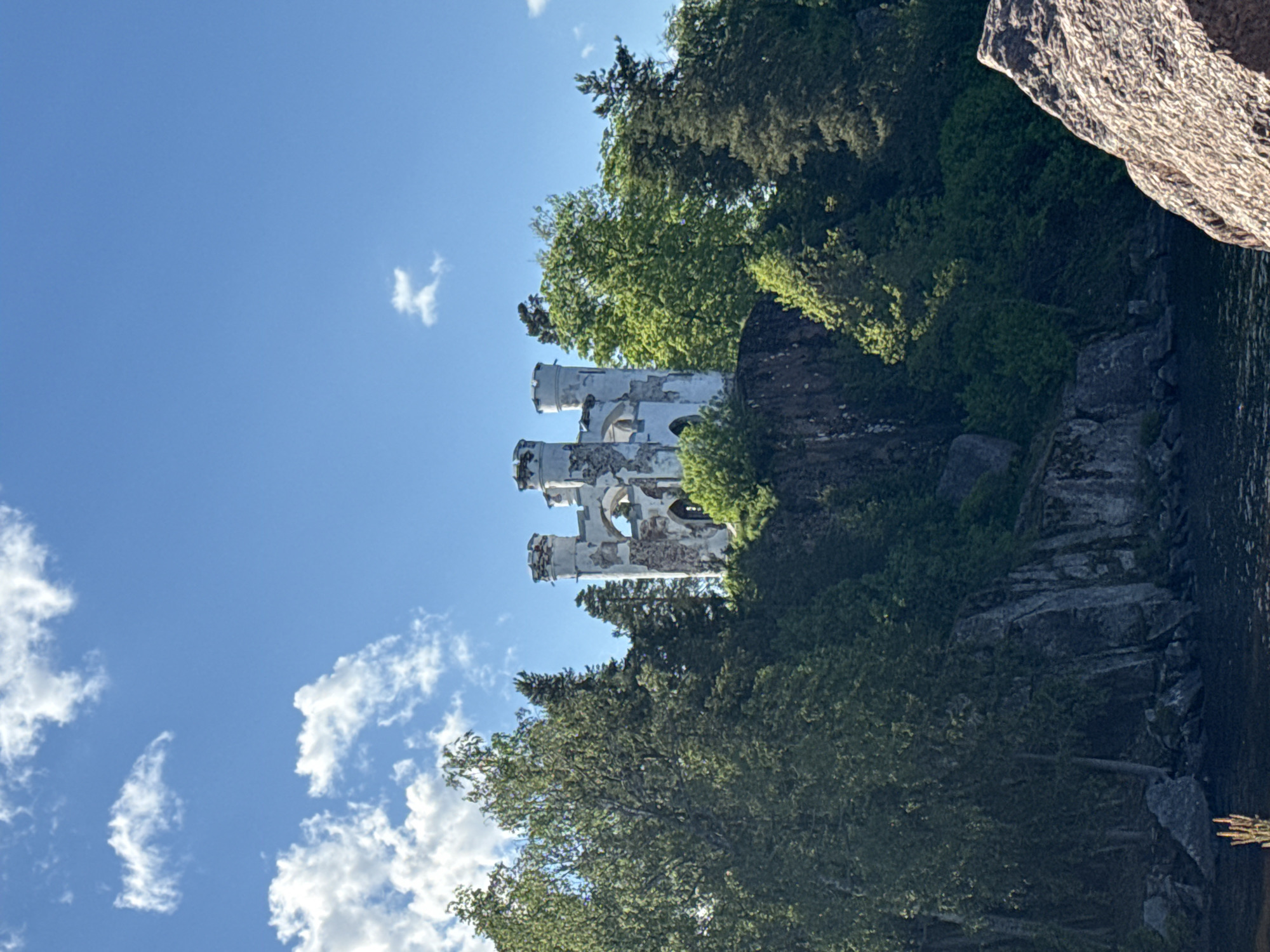 The image depicts the ruins of the St. Hyacinth Fort in Vyborg, Russia. The ruined structure, with its weathered, white towers, is partially obscured by lush green trees and rocky terrain. A body of water is visible on the right, potentially a part of the Gulf of Finland. The sky above is blue with scattered white clouds, indicating fair weather. A large rock is visible in the right corner of the image.