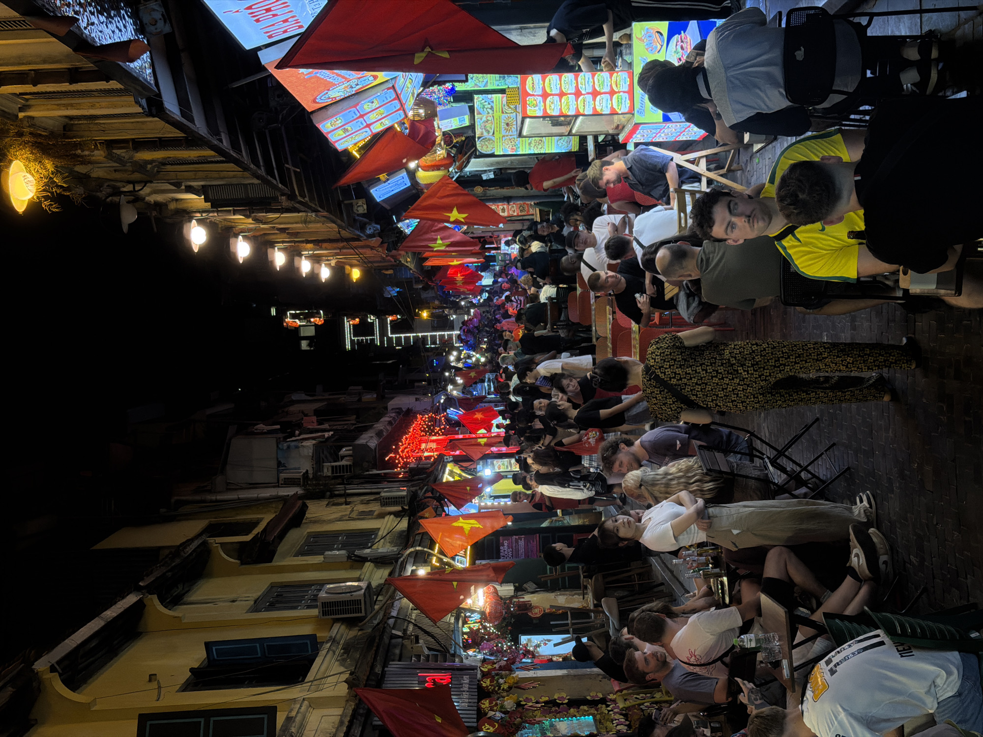 This vibrant nighttime photograph depicts a bustling street scene in Hanoi, Vietnam, filled with people socializing and dining outdoors. The narrow street stretches into the distance, illuminated by a warm glow from string lights, lanterns, and various glowing signs.
Numerous individuals are seated at small, low tables and chairs, characteristic of Vietnamese street food culture, which line both sides of the thoroughfare. People, appearing to be a mix of locals and tourists, are engaged in lively conversation, eating, and drinking, contributing to a relaxed and social atmosphere. Many faces are visible, some looking directly towards the camera, others focused on their companions.
Overhead, a series of red Vietnamese flags, featuring a yellow star, are strung across the street, adding a distinct cultural element. Further down the street, additional red lanterns and festive decorations enhance the evening ambiance. The buildings flanking the street are multistory structures, some displaying traditional architectural details and equipped with air conditioning units. Illuminated signs advertise establishments, with the phrase "BIA HƠI" clearly visible on a prominent sign, indicating a popular local draft beer spot. Other signs show text like "PHO" and "MENU," suggesting the array of food and drink available. The scene captures a quintessential lively evening in Hanoi, alive with human connection and urban energy.