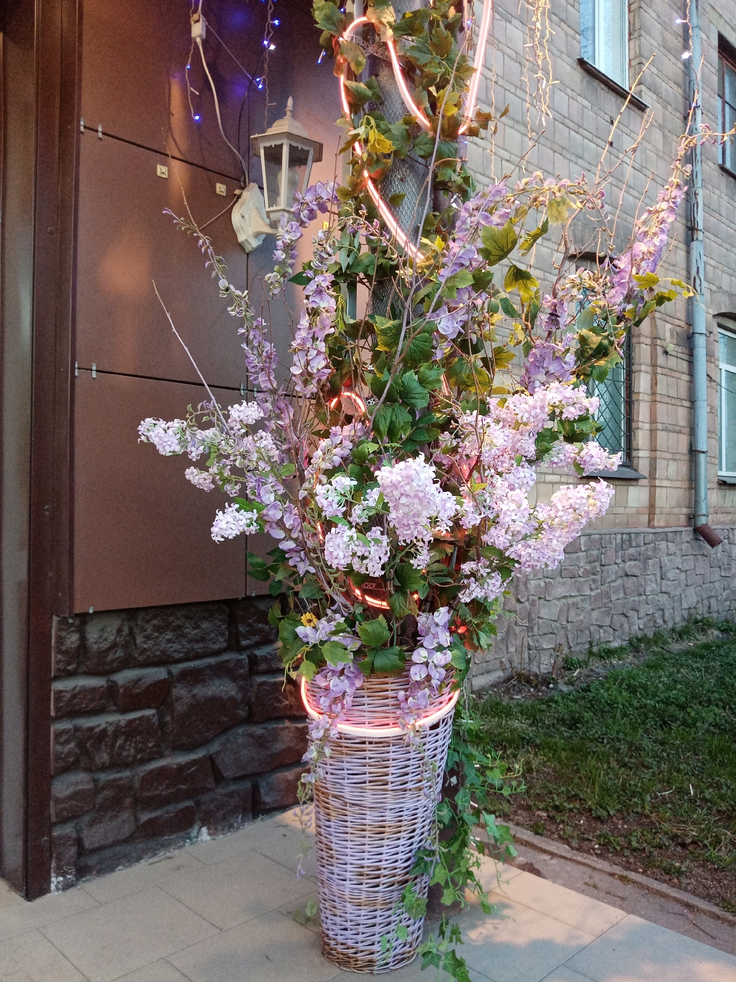 The image shows a tall, cylindrical wicker basket overflowing with artificial purple and white flowers and greenery, illuminated by a neon light that wraps around the arrangement. The basket sits on a light-colored tiled sidewalk. Behind the basket are two buildings, one with brown siding and a stone base, the other a light gray brick building with windows. A streetlamp is mounted on the brown building. The photograph was taken in Elektrostal, Russia.