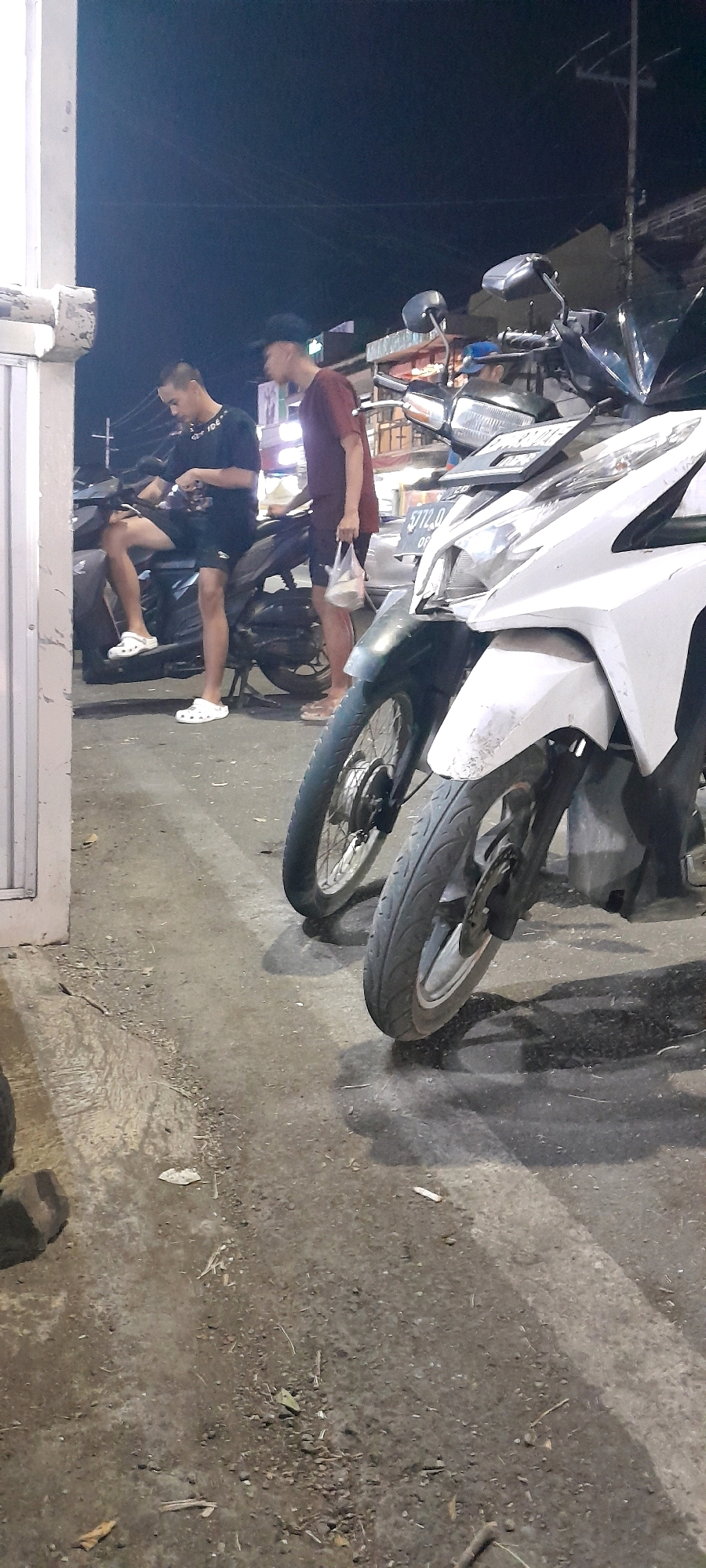 Two young men are present in the image, which appears to be taken at night on a street in Rogojampi, Indonesia. The man on the left is seated on a dark-colored motorcycle, wearing a black t-shirt and shorts, with white clogs on his feet. He is looking down at his phone. The man on the right is standing next to the motorcycle, wearing a maroon t-shirt and shorts, and holding a plastic bag in his right hand. He is looking towards the left.
Several motorcycles are parked in the scene. In the foreground, a white motorcycle is partially visible, with its front wheel and lower fairing in focus. Behind it, a dark-colored motorcycle is more fully visible, with both wheels and the frame discernible. The lighting suggests an outdoor environment, possibly a street or a parking area, with artificial light sources illuminating the scene. The ground is paved with concrete and appears somewhat dirty, with debris scattered on the surface. In the background, buildings and streetlights are faintly visible, indicating an urban or semi-urban setting. The overall impression is of a casual, everyday scene taking place in a Southeast Asian locale at night.