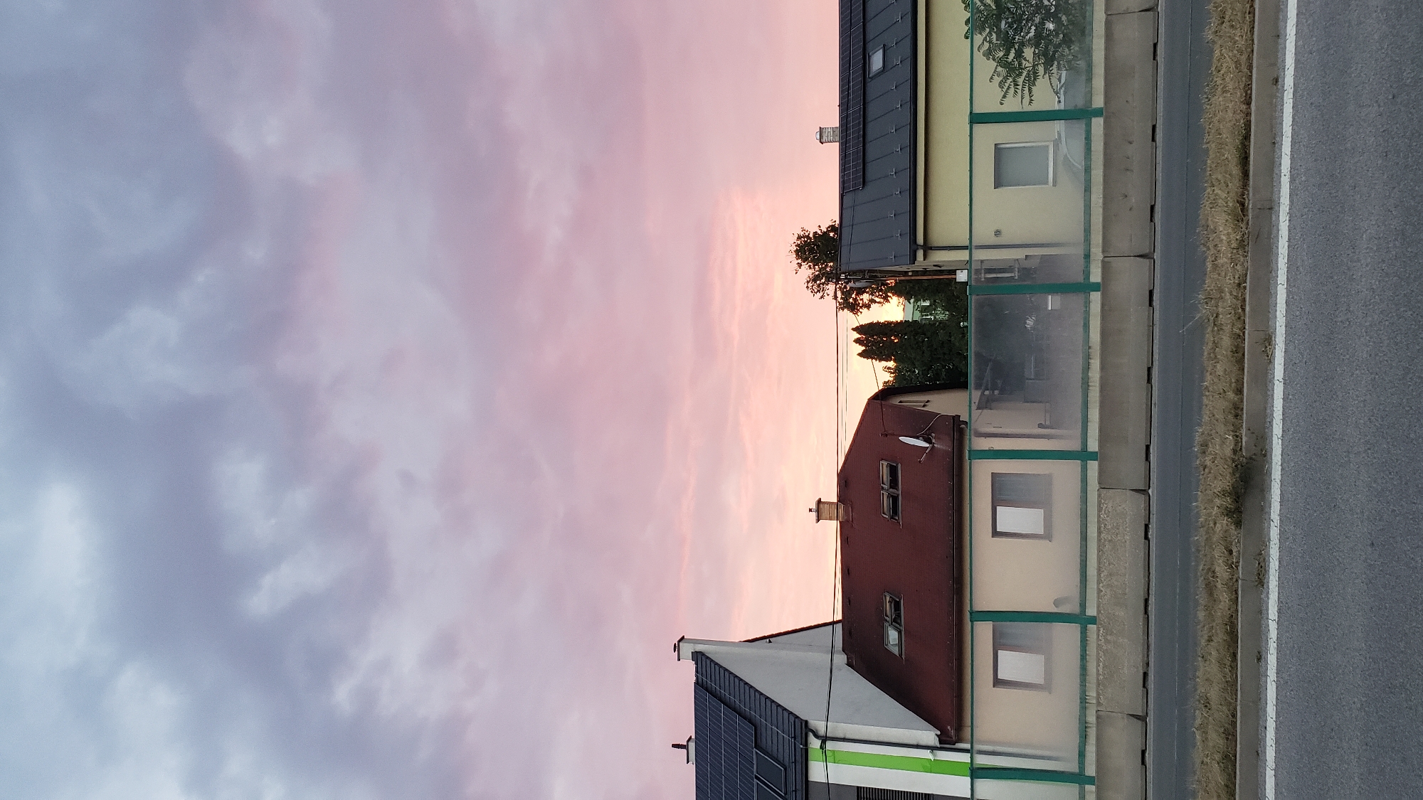 The image depicts a row of houses behind a sound barrier, with a street in the foreground. The sky is filled with clouds tinged with pink and purple hues, suggesting either dawn or dusk. The houses have various roof types, including solar panels and traditional red tiles. Some windows are visible, as is a satellite dish on one of the houses. The sound barrier is made of concrete and transparent panels. There are no people visible in the image. The overall scene is calm and quiet, with the soft colors of the sky dominating the upper portion of the frame. The location is the city of Prešov, Slovakia, likely on the outskirts where residential areas meet transportation routes.