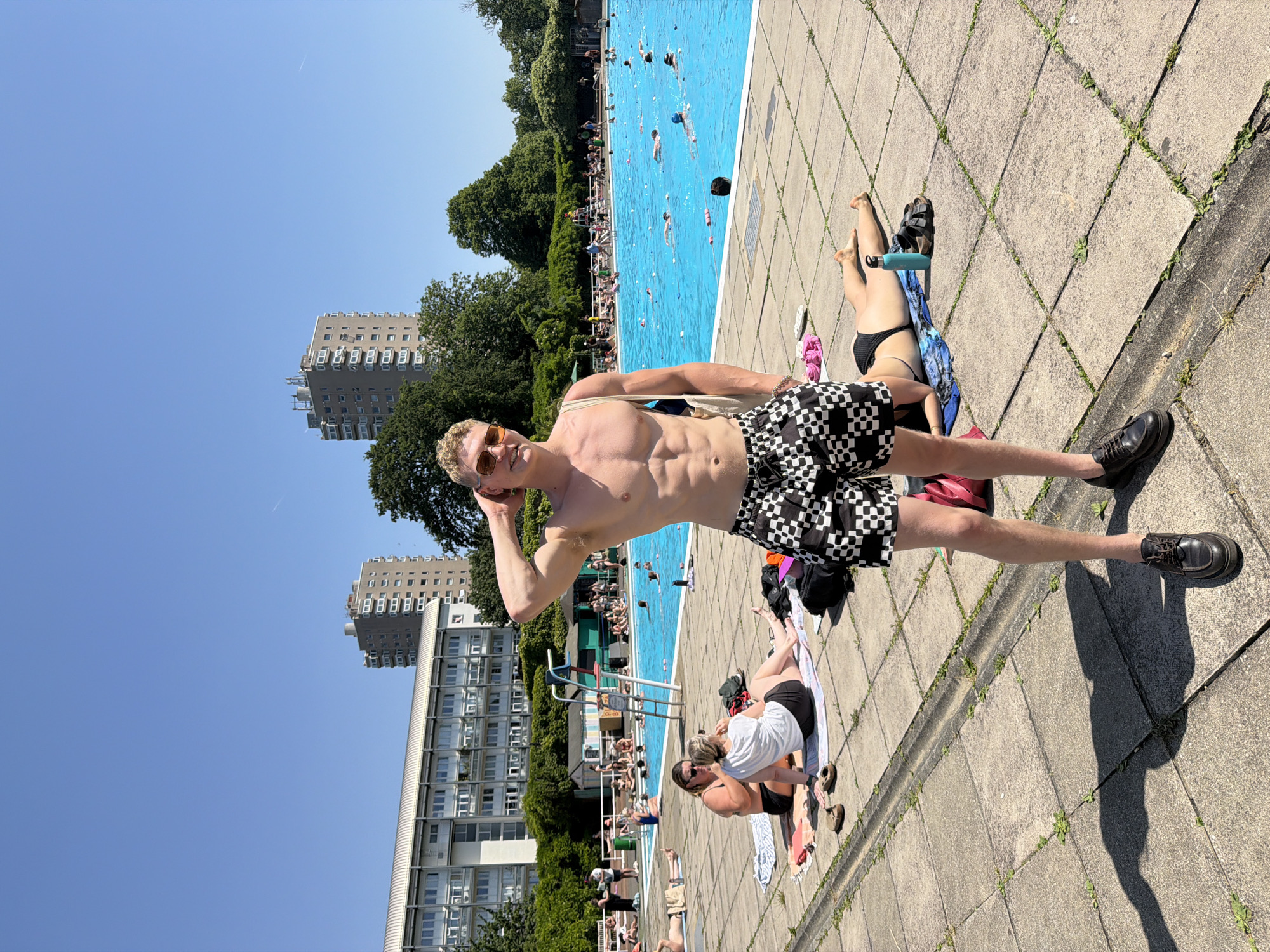 A vertically-oriented photograph shows a man in checkered shorts, shirtless and wearing sunglasses, striking a pose beside an outdoor swimming pool on a sunny day. The man is blond and appears to be in his twenties.
The pool is rectangular and filled with blue water, with people swimming and playing in it. A lifeguard stand is visible near the edge of the pool. Sunbathers lie on the paved area surrounding the pool. In the background, multiple high-rise buildings are visible against a clear blue sky. Green trees are interspersed between the buildings.
The image was most likely taken in London, United Kingdom.