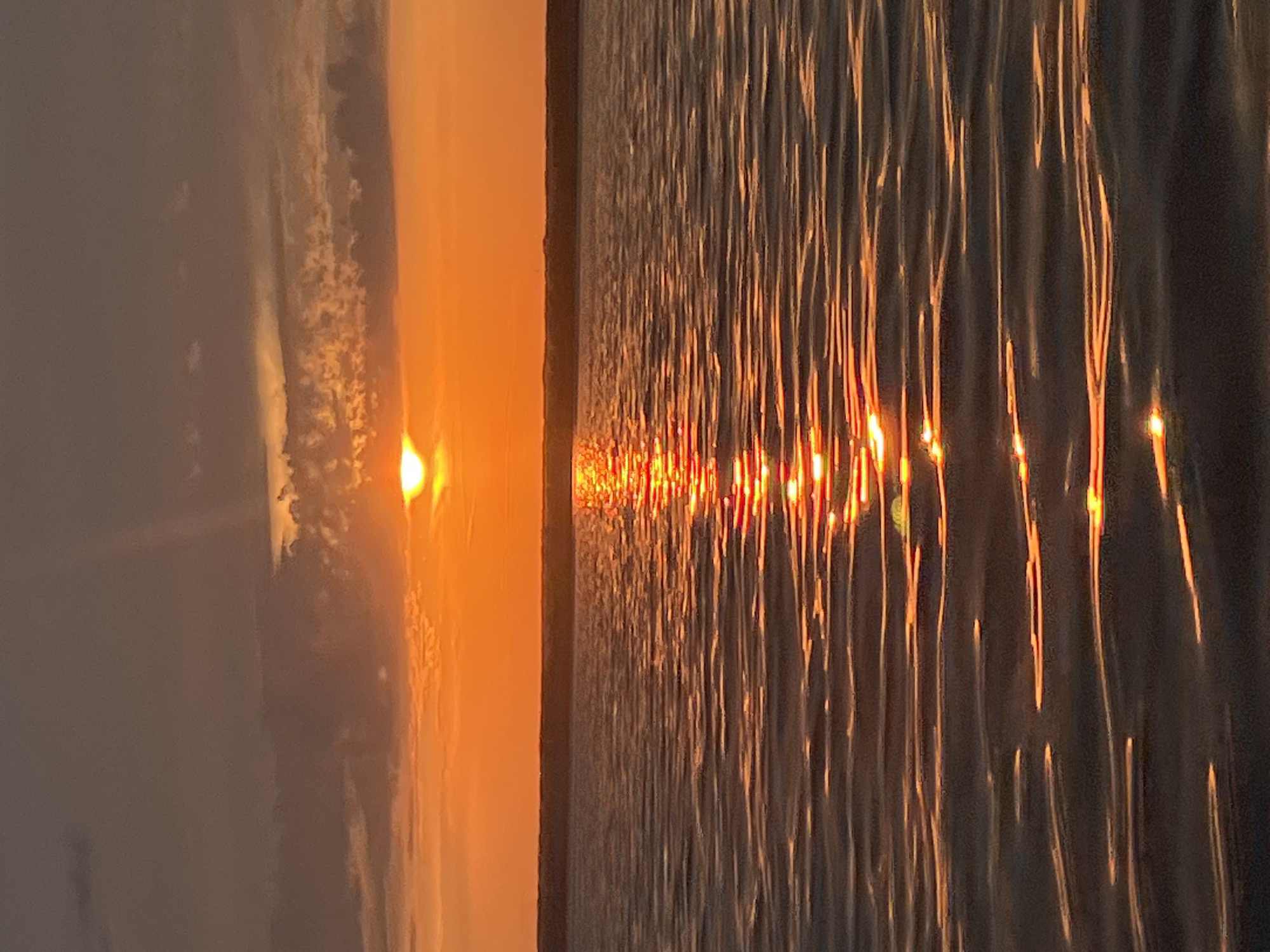 A sunset over the sea in Beregovoe, Russia, is captured in this vertical shot. The sun, a bright orb, is partially visible above the horizon, casting a vibrant orange glow across the sky and the water. Clouds, tinged with the same orange hue, dot the dusky sky. The surface of the sea is rippled, reflecting the fiery colors of the sunset in a shimmering path that stretches towards the viewer. The overall atmosphere is serene and tranquil, evoking the beauty of nature at the close of day.