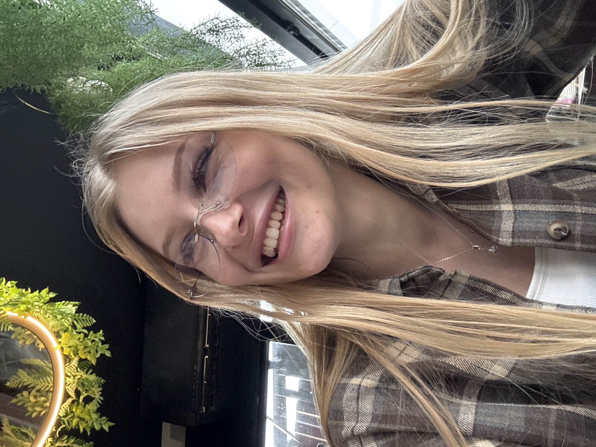 A young woman with fair skin and blonde hair is smiling at the camera while wearing glasses and a plaid shirt. She appears to be indoors, possibly in a restaurant or cafe, as greenery and a window are visible in the background. The location cannot be determined to be Perm, Russia based solely on the image.