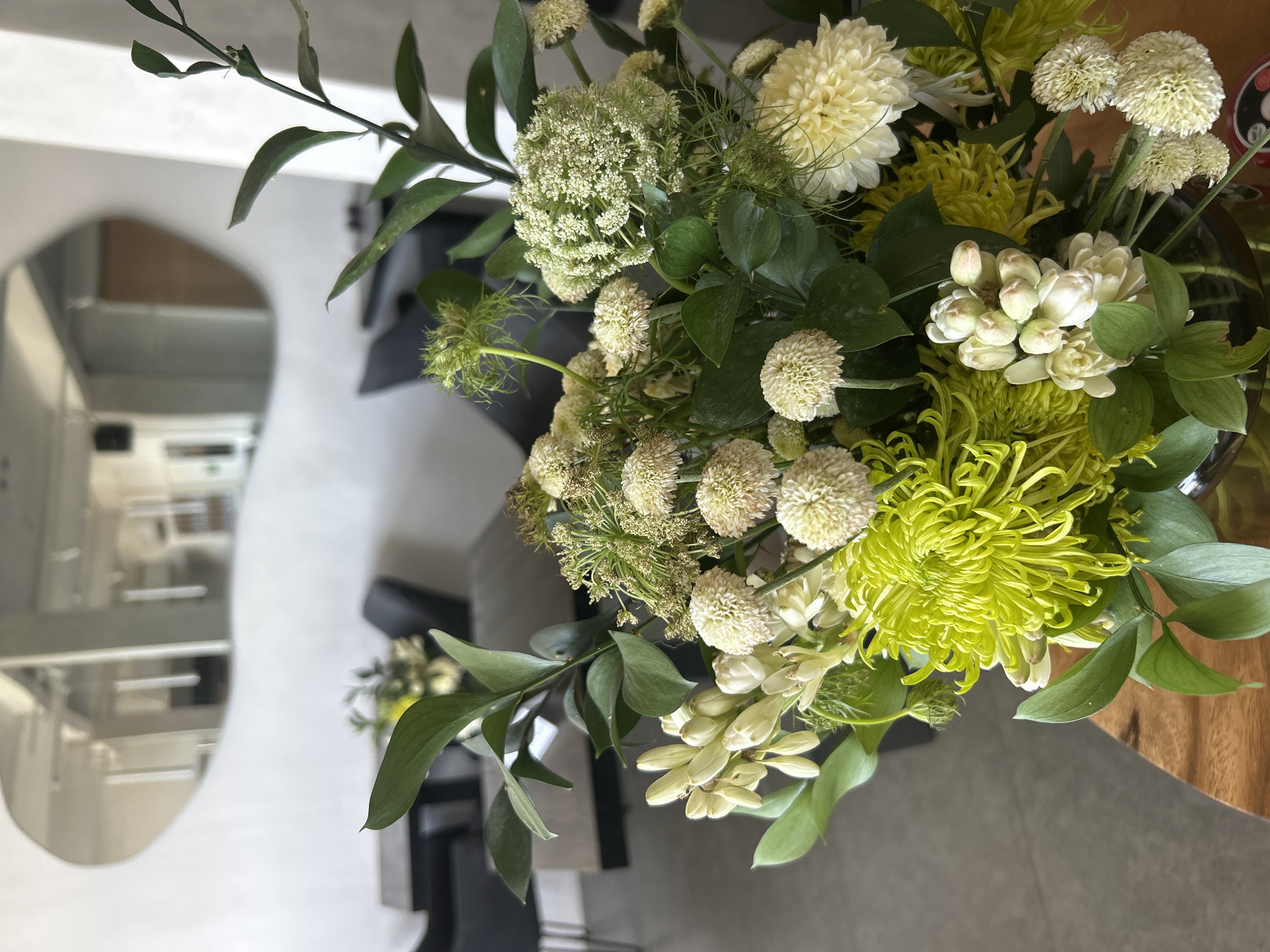 A floral arrangement with white and green flowers sits on a wooden surface in a room in Kuta Selatan, Indonesia. The flowers include chrysanthemums, queen anne's lace, craspedia, and ruscus. The background shows a salon with chairs and a mirror.