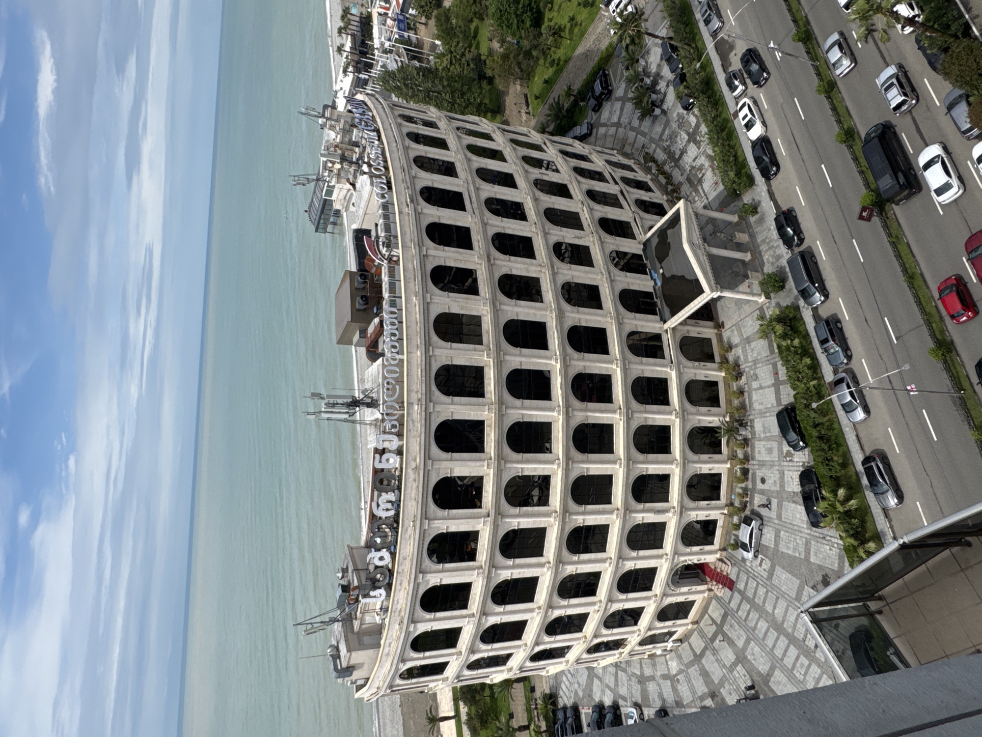 This is an elevated view of the Colosseum Marina Hotel in Batumi, Georgia. The distinctive, white, semi-circular building is situated adjacent to the Black Sea and a multi-lane road with moderate car traffic. The facade of the building mimics the arches of the Roman Colosseum. The area around the hotel features patterned pavement, greenery, and palm trees. The sky is partly cloudy, with the sea extending toward the horizon.