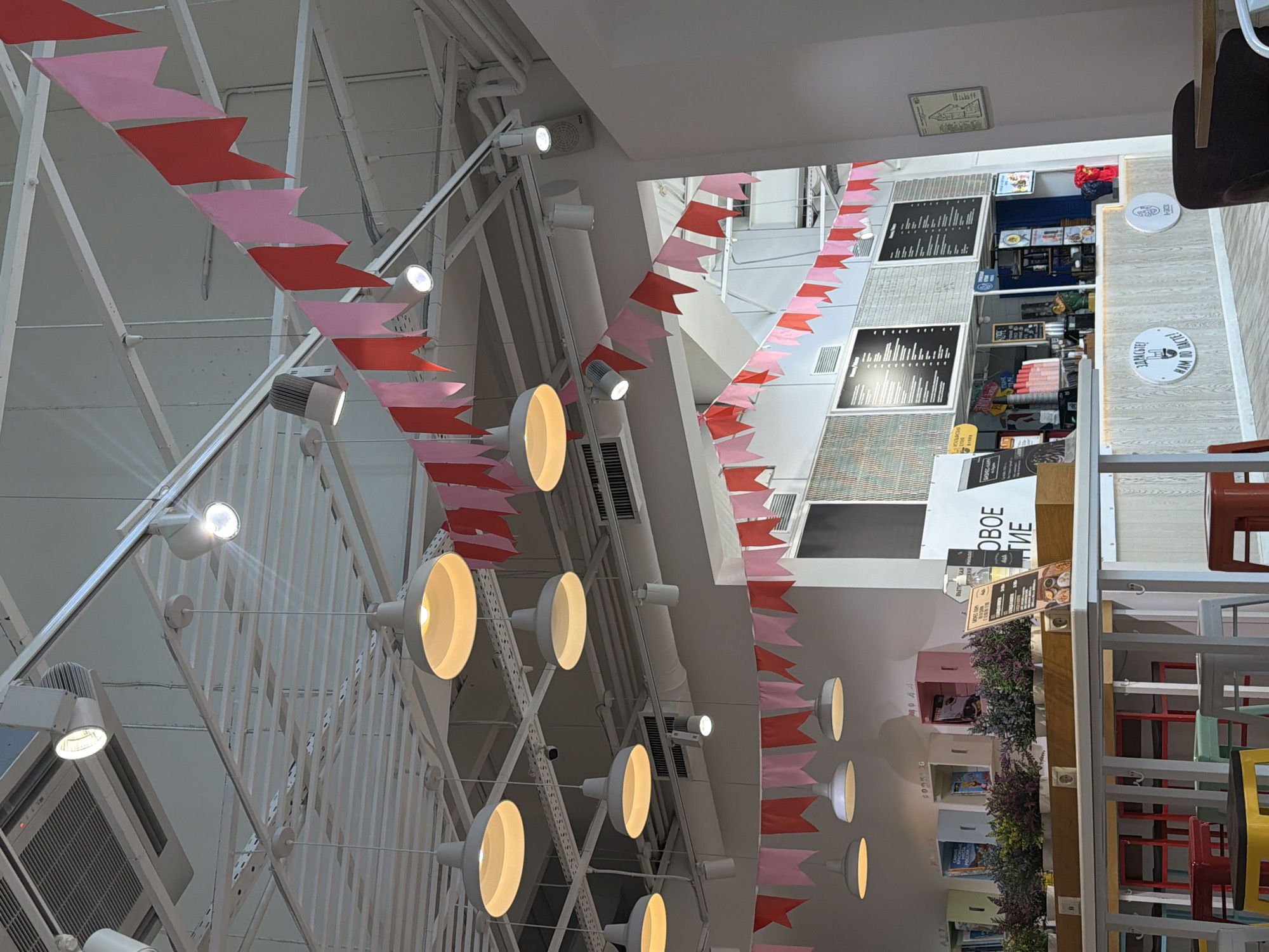 The image shows the interior of a brightly lit commercial space, likely a cafe or restaurant, in Lyubertsy, Russia. The ceiling features a complex structure of white beams and metal supports, adorned with rows of decorative pink and red pennant flags. Rows of industrial-style light fixtures are attached to the ceiling's framework.
In the background, elements of a food service establishment are visible, including menus, serving counters, and seating areas. The decor includes plant displays and shelving filled with books. The space is modern and appears well-maintained.