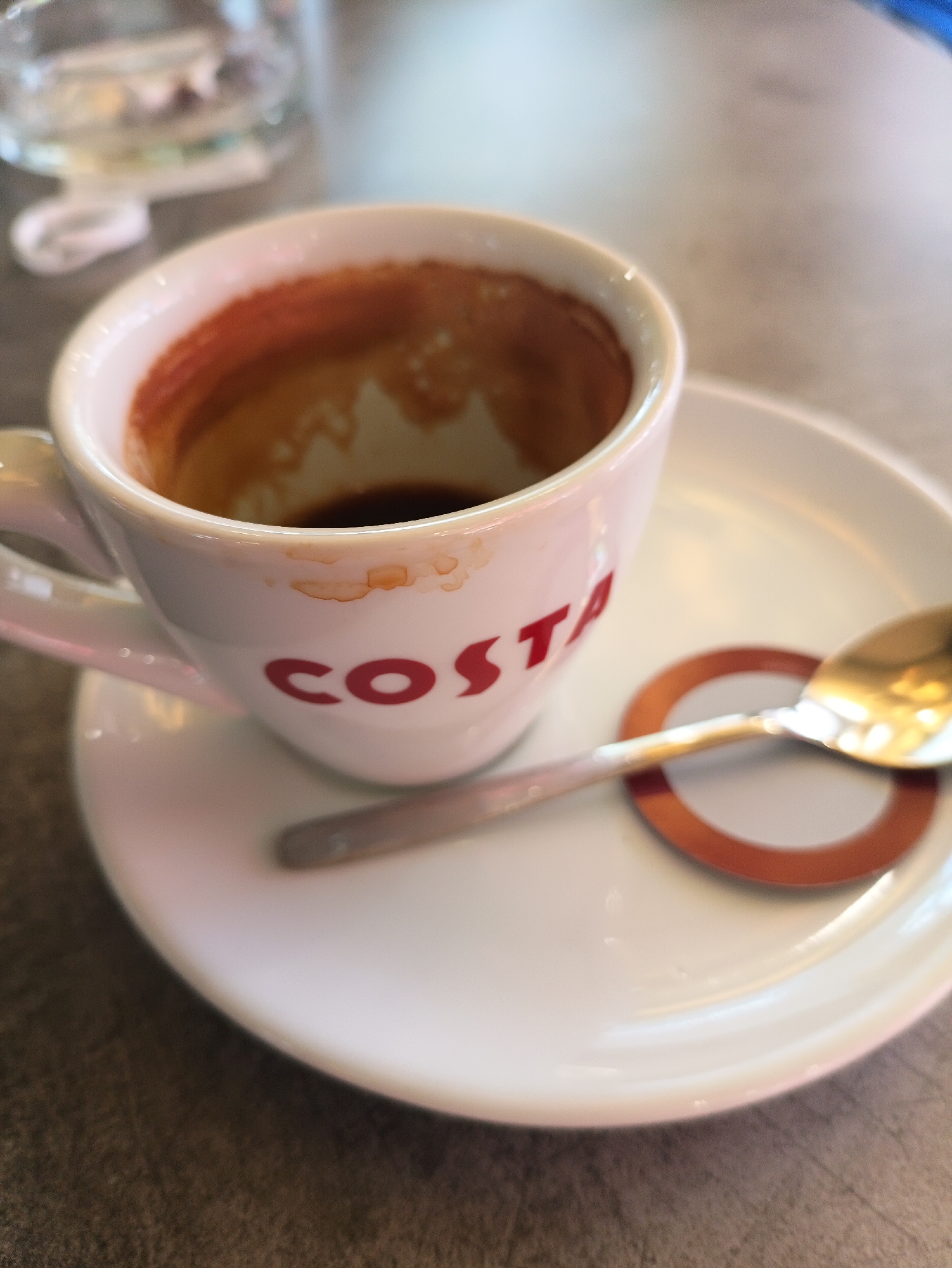 The image shows an almost-empty white ceramic Costa coffee cup on a white saucer. A gold-colored spoon rests on the saucer near a small ring with a white interior and brown exterior. The cup has the word "COSTA" printed on it in red. Coffee residue is visible inside the cup and around its rim. A blurry glass of water is visible in the background. The surface is a dark gray color. The image does not contain any explicit information about the location being Karnobat, Bulgaria.