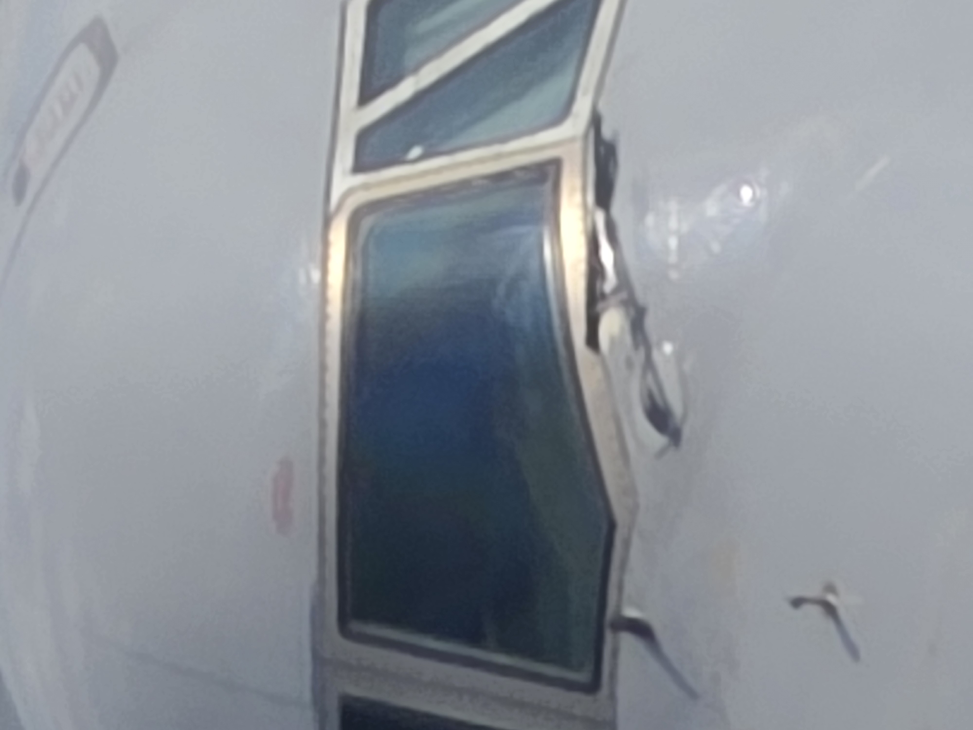 The image shows the front section of an airplane, likely a passenger jet, as seen from a low-angle perspective. The focus is on the cockpit windows, which are dark and reflective, showing a hint of the sky and clouds. The aircraft's fuselage is a light, neutral color, possibly white or silver, with some subtle reflections of light. The overall impression is of an aircraft ready for flight or recently landed, with no people or specific external environment details visible within the frame. There is no information within the image that allows for determining the location in the city of Frankfurt, Germany.