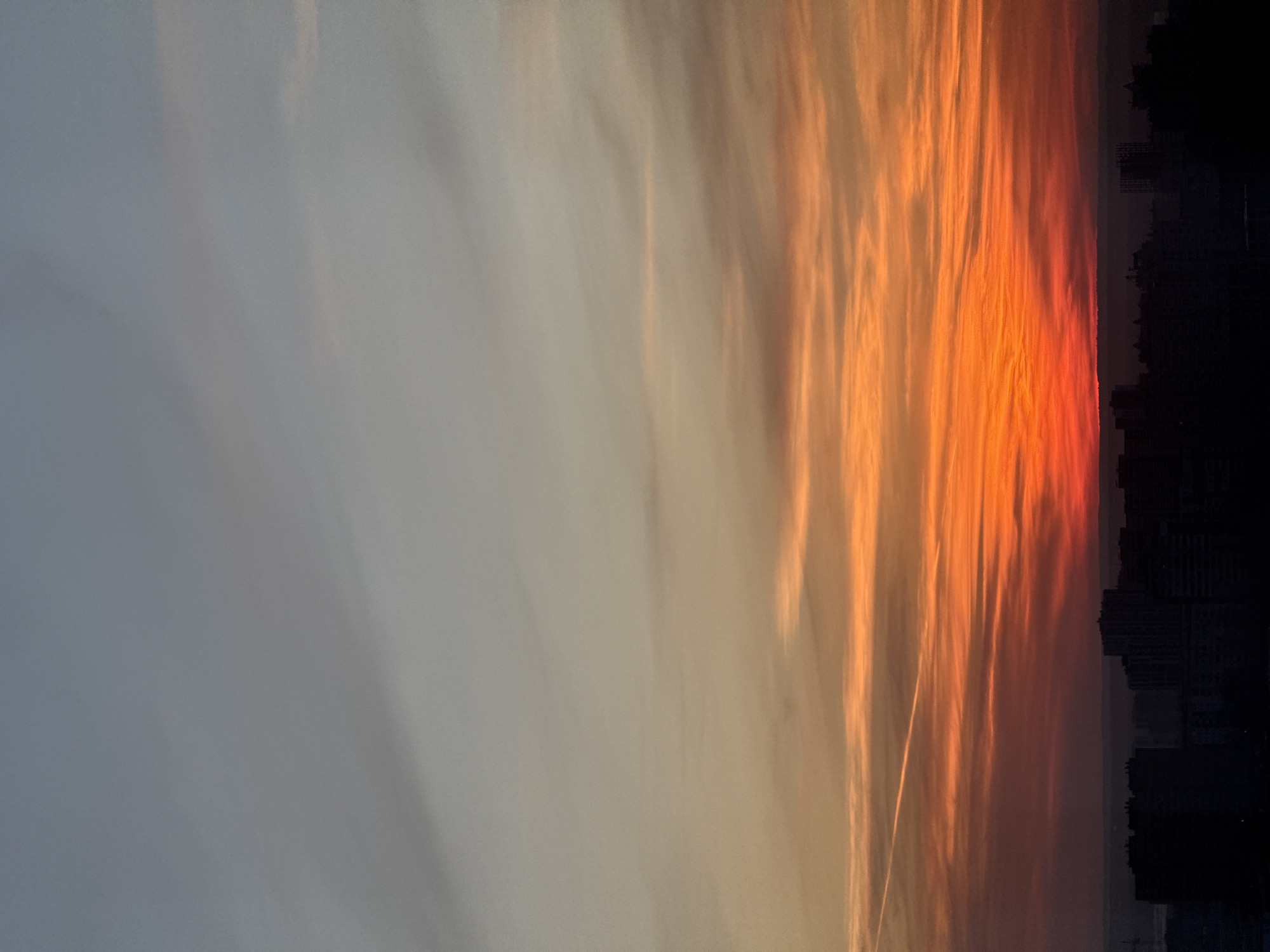 The image displays an urban sunset or sunrise over the city of Perm, Russia. The sky occupies the majority of the frame, exhibiting a gradient from light blue-grey at the top, transitioning through muted orange and peach tones, to intense orange and deep red at the horizon. Horizontal streaky clouds are prominent across the middle and lower sky, illuminated by the low sun. A single, thin, white contrail from an aircraft extends diagonally from the upper left towards the lower right within the orange-red portion of the sky. The lower portion of the image reveals a dark, silhouetted cityscape, comprising numerous buildings of varying heights. These structures are indistinct due to the lack of direct light, appearing as solid black forms against the bright sky. No people, discernible actions, or interactions are visible within the scene. The environment is an elevated panoramic view of an urban landscape at either dawn or dusk.