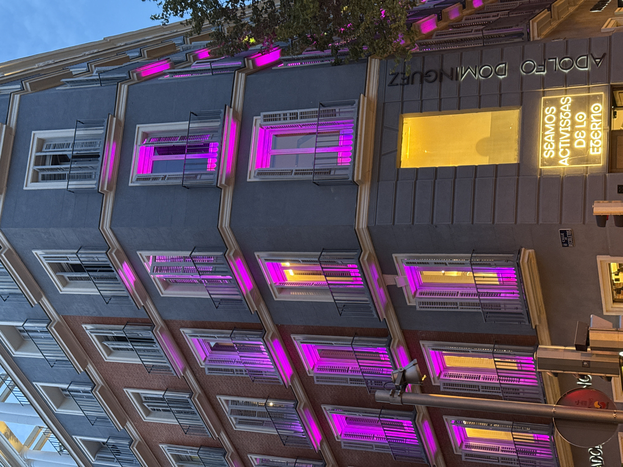 A low-angle, eye-level outdoor shot of a tall building in Madrid, Spain, shows illuminated windows, some with a vibrant pink or purple glow, others with a golden yellow light. The building has a grey facade with alternating sections of exposed brick. The name "ADOLFO DOMINGUEZ" is illuminated in a white sans-serif typeface on the upper part of the building, and another sign beside it shows the words "SEAMOS ACTIVISTAS DE LO EBERNO" in a glowing yellow hue. Metal window grates extend from the window frames. A street sign with the word "RECOLETOS" is visible. A street lamp, and the partial red glow from another sign, appear on the bottom right, with branches from a tree reaching into the upper right. The sky in the background is a deep blue.