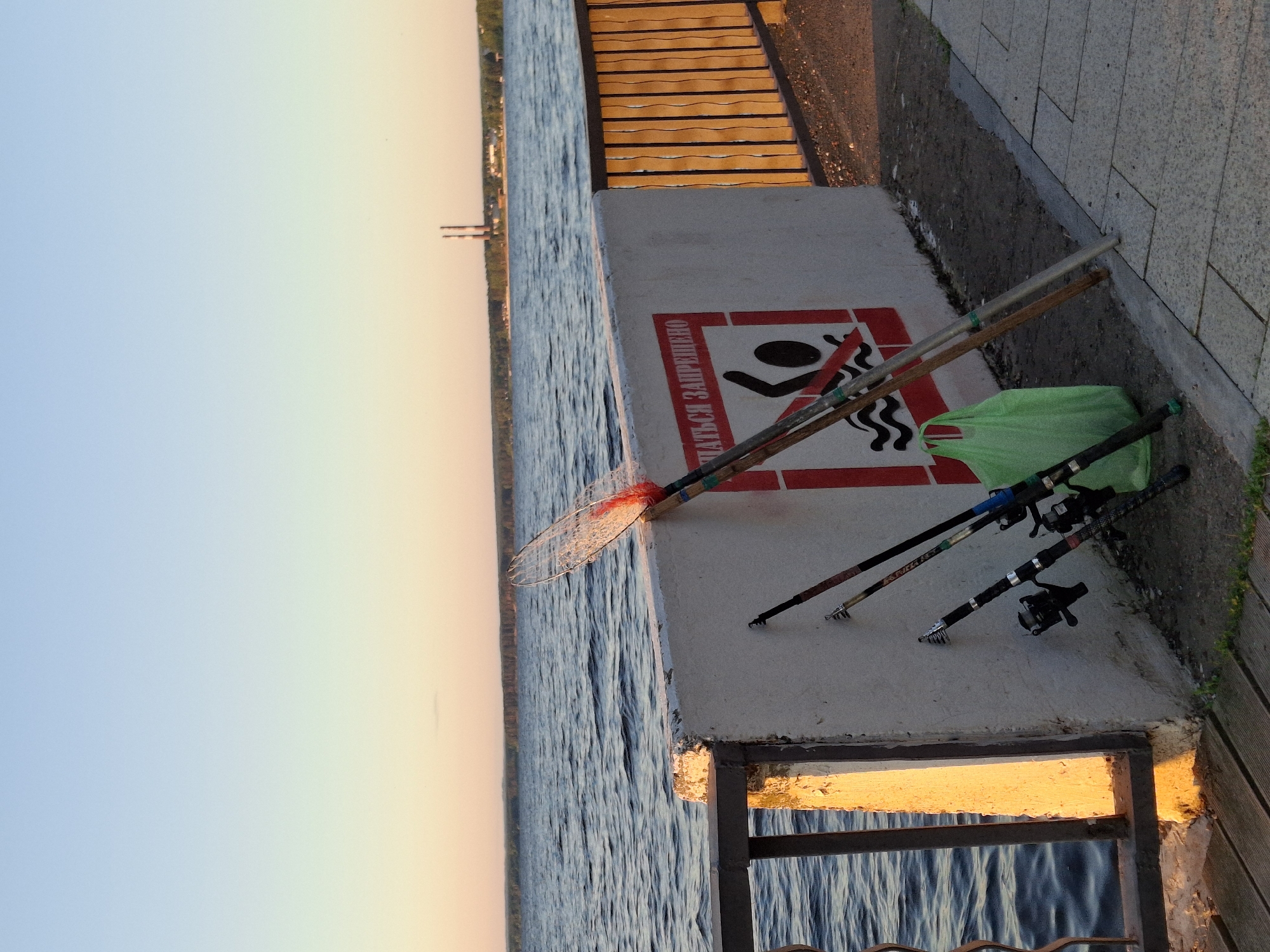 The image depicts a serene waterside scene in Chaikovsky, Russia. The sun is setting, casting a warm, golden glow on the environment, with the sky transitioning from a soft blue to a pale orange. The foreground features a concrete embankment overlooking a body of water with gentle ripples.
Several fishing rods are propped up on the embankment. One fishing net with a red handle is positioned nearby. A bright green plastic bag lies on the ground next to the fishing gear. A prominent sign with a red border is painted on the concrete, featuring a pictogram of a person falling into water, accompanied by the Russian text "КУПАТЬСЯ ЗАПРЕЩЕНО" (KUPAT'SYA ZAPRESHCHENY), which translates to "SWIMMING IS PROHIBITED".
In the background, a wooden boardwalk or pier with horizontal planks is visible, leading away from the embankment. The overall atmosphere is peaceful and suggests a quiet evening by the water, likely a popular spot for fishing, despite the prohibition against swimming. The natural light and the presence of fishing equipment indicate a moment of pause after a day's activity or in anticipation of fishing.