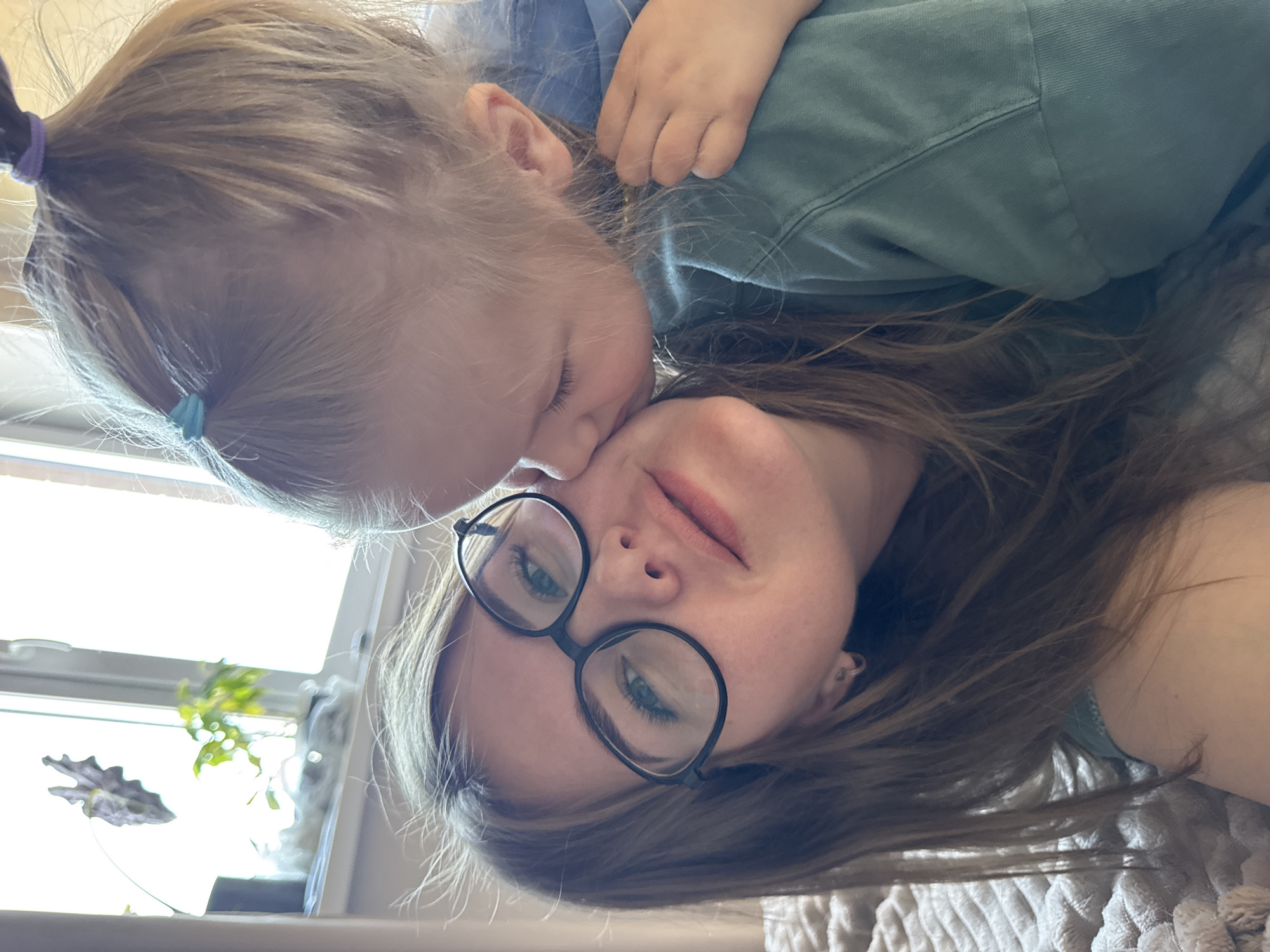 A photograph shows a light-skinned woman wearing black-framed glasses with a light-skinned toddler nestled against her face. The toddler, positioned at the top of the image, has light blonde hair pulled back into two small pigtails with a light blue hair tie visible. The child wears a light blue top and is in the process of either kissing or nuzzling the woman's cheek, close to her glasses. The woman has medium-length light brown hair and is wearing a light-colored top. The background shows a window with a plant visible on the windowsill. The scene is indoors, and the perspective suggests that the photograph was taken from above. There is no specific information provided to suggest that this photograph was taken in Ферма, Russia.