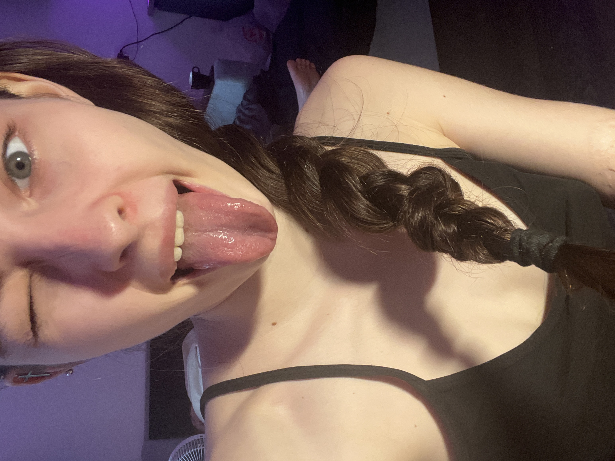 The image shows a person from a close, angled perspective. The person has fair skin, brown hair styled in a braid, and is wearing a black tank top. The person is sticking their tongue out. The background is dimly lit, with hints of furniture and a purple hue suggesting artificial lighting. The location in Saint Petersburg, Russia, cannot be determined from this image.