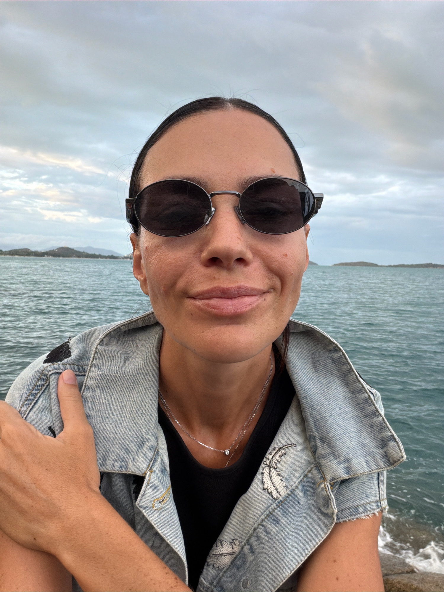 A woman is photographed from the chest up, facing the camera, with a serene expression and wearing sunglasses. She is dressed in a light blue denim jacket over a black top, with a delicate silver necklace around her neck. Her left arm is bent and resting on her chest. The background features a tranquil body of water, likely the sea, with distant, low-lying islands and a cloudy sky overhead. The scene suggests a coastal environment, possibly a pier or a rocky shoreline, characteristic of areas like Ko Samui District, Thailand, known for its beautiful beaches and offshore islands. The lighting is diffused, indicating an overcast day or early evening.