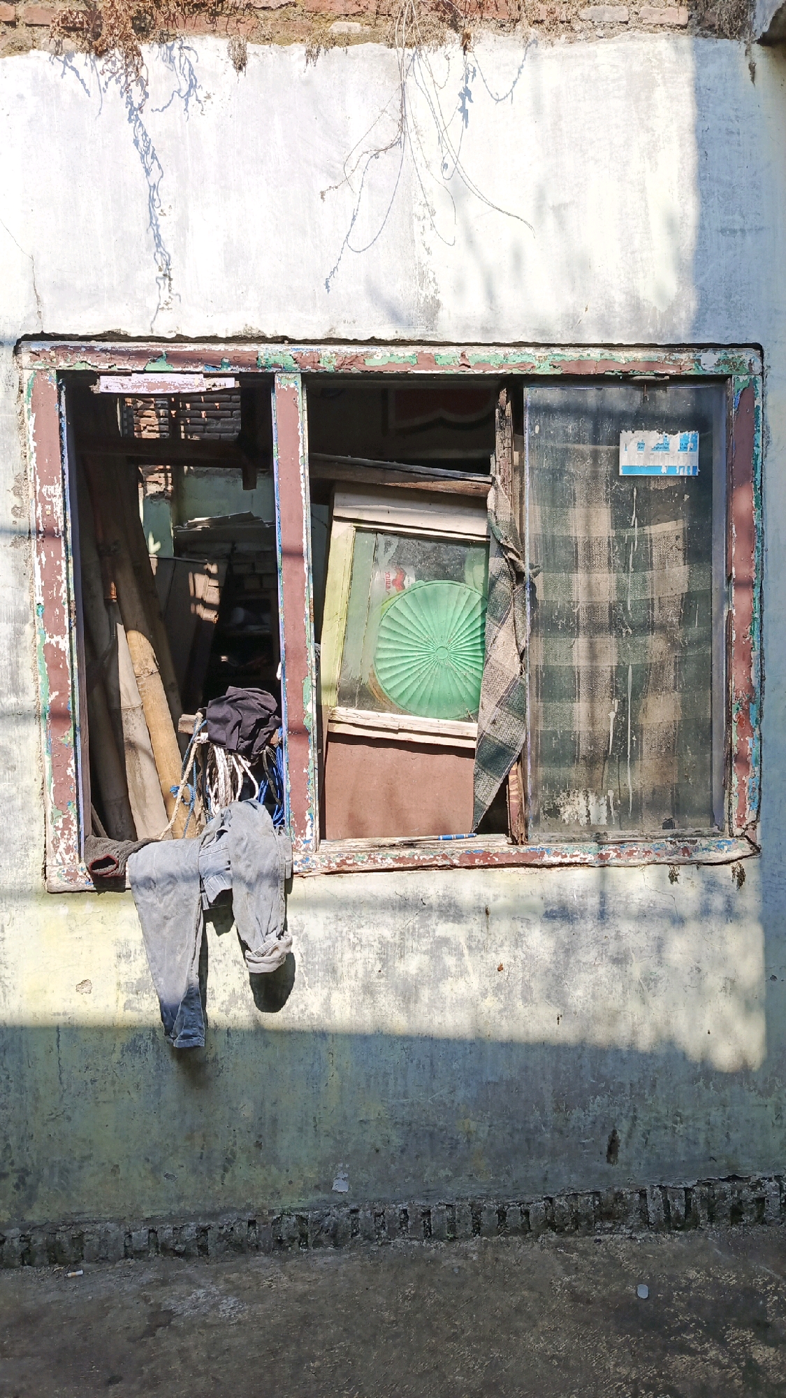 This image depicts a weathered and somewhat dilapidated window of a building in Pekauman Kulon, Indonesia. The window frame is aged, with peeling paint in shades of red and green. The left side of the window is open and reveals a cluttered interior containing several lengths of bamboo, a black duffel bag, and some rope. A pair of grey trousers is draped over the lower part of the window frame, with the legs hanging down. The right side of the window is covered by a glass pane that is smudged and dirty, with a checkered fabric curtain hanging loosely over it. A green, circular object, possibly a fan or decorative item, is visible behind the glass. The wall surrounding the window is textured and discolored, with patches of growth clinging to the upper edge. Sunlight casts shadows on the wall and the ground below, suggesting an outdoor setting. The ground in front of the building is paved with rough concrete and brickwork.