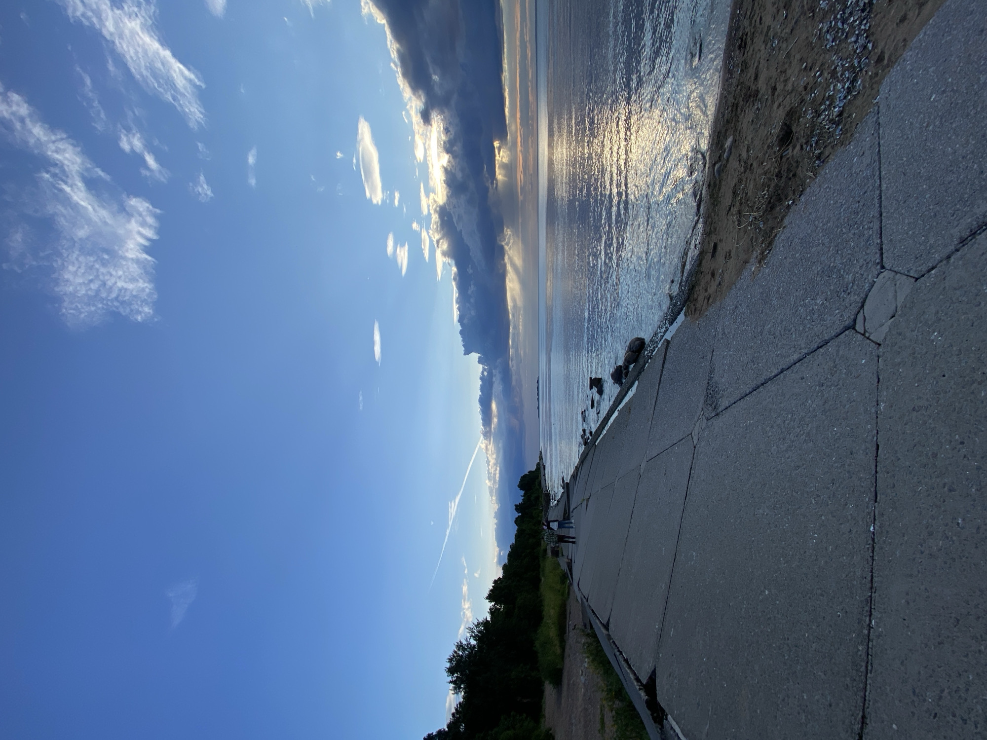 The image depicts a scenic view of the Gulf of Finland coastline in Sestroretsk, Russia, during what appears to be sunset. A concrete embankment, showing signs of cracking and disrepair, runs along the left side of the frame, leading the eye towards the expansive body of water. Two individuals, a man and a woman, are seen walking side-by-side on the embankment, their backs to the camera.
The sea is calm, with gentle ripples reflecting the soft hues of the sky. A bank of dark clouds is positioned over the horizon, partially obscuring the sun, but allowing dramatic rays of light to pierce through and illuminate the water. A faint contrail from an aircraft stretches across the bright blue sky above.
The shoreline to the right of the embankment is a mix of sand and small stones, with scattered shells visible. Lush green trees and foliage line the embankment on the left, creating a natural border against the urban landscape. The overall atmosphere is serene and tranquil, capturing a moment of natural beauty at dusk.