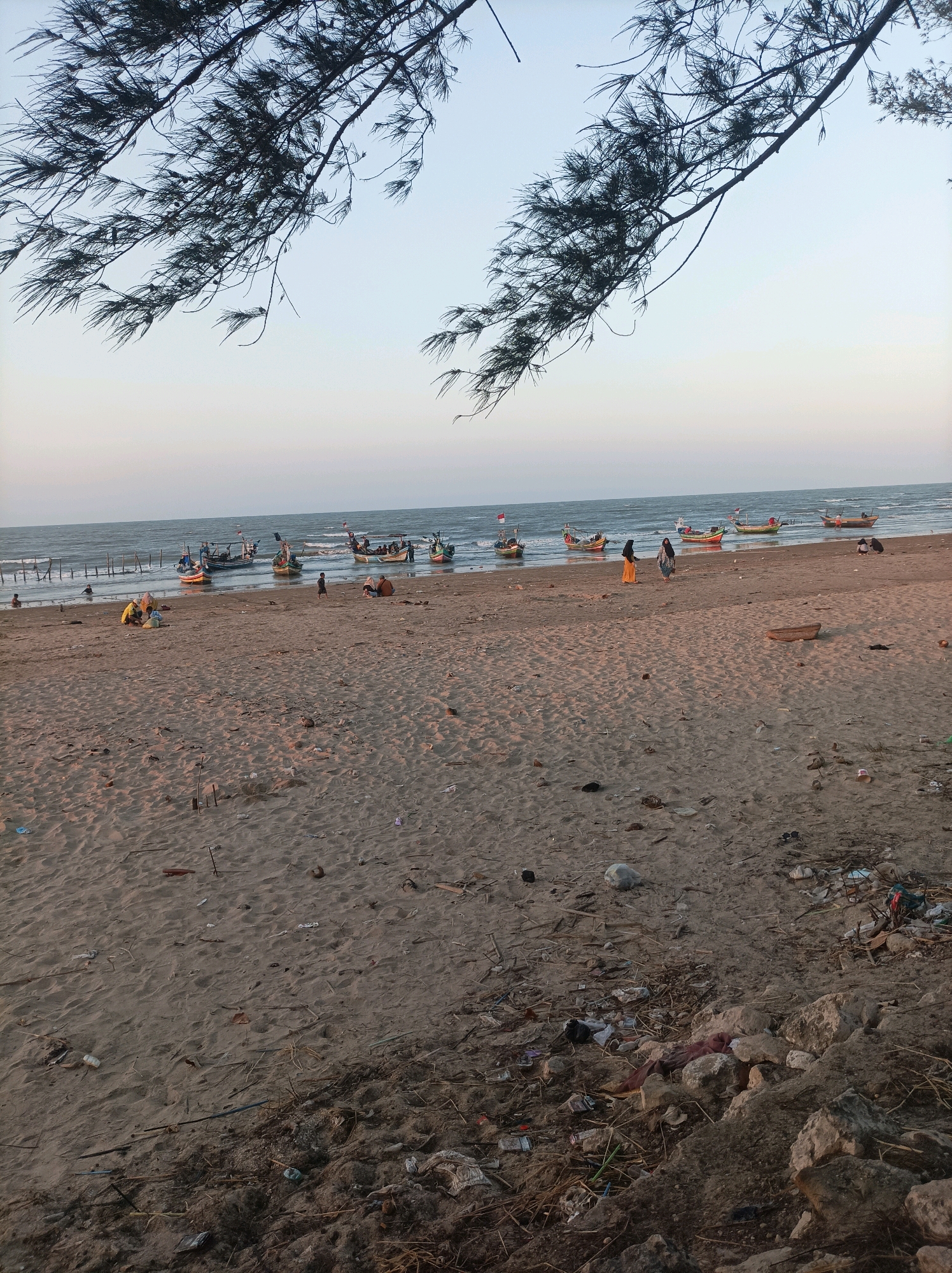 The image shows a sandy beach in Palang, Indonesia, with several fishing boats anchored offshore. The sky is a pale blue, and the ocean is calm. The beach is littered with various debris, including plastic bottles, bags, and dried leaves. In the foreground, there are a few people sitting and walking on the sand. The trees in the upper portion of the image overhang the beach, casting shadows on the sand. The scene depicts a typical day at a fishing village, with a mix of human activity and natural elements.
