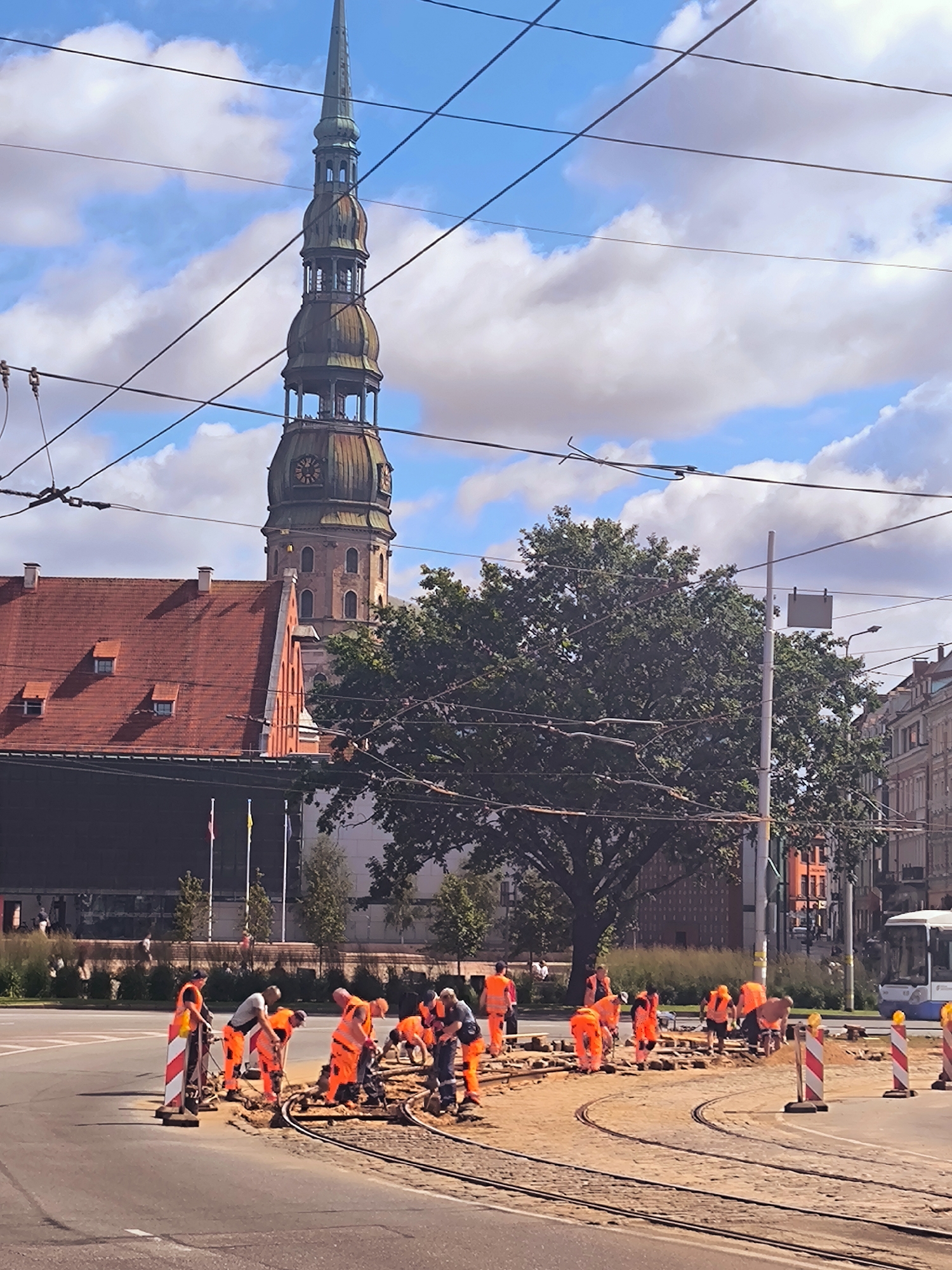 The image captures a street scene in Riga, Latvia, specifically in front of the House of the Blackheads and St. Peter's Church. A group of construction workers in bright orange safety vests and trousers are actively engaged in repairing tram tracks. They are spread across the work area, some bending down to handle tools and materials, while others are standing and observing. The ground around the tram tracks is dug up and covered with sand and gravel, indicating ongoing construction. In the background, the iconic spire of St. Peter's Church, with its distinctive green-topped architecture and clock face, dominates the skyline. Adjacent to the church is a building with a red tiled roof and multiple chimneys. Lush green trees partially obscure the view of the buildings, suggesting a well-treed urban environment. Overhead, a complex network of tram wires crisscrosses the sky, typical of Riga's extensive tram system. A white bus is visible in the far right background, and buildings line the street on the right side of the image. The sky is a mix of blue with scattered white clouds.