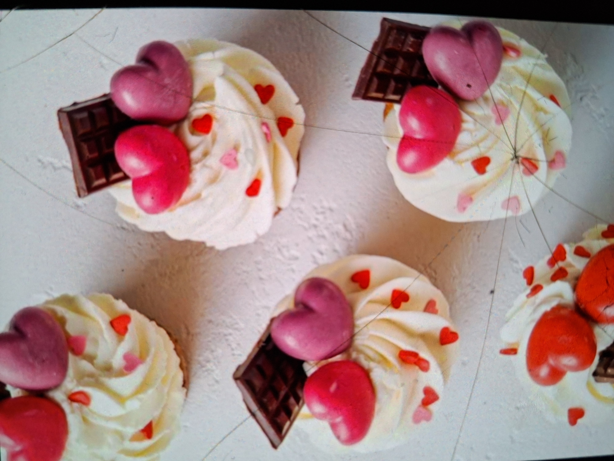 This is a close-up, overhead shot of several cupcakes on a white, textured surface, likely a table. The cupcakes are decorated with swirls of white frosting, red and pink heart-shaped sprinkles, and some are topped with chocolate pieces and larger, heart-shaped candies. The heart candies appear to be a light pink or purple and a bright pink or red. The chocolate pieces are dark brown, segmented pieces. The scene is well-lit, and the focus is sharp on the cupcakes. There is no visible text in the image. The setting suggests a celebratory occasion, possibly Valentine's Day, given the heart motifs. The overall mood is festive and sweet. The image appears to be taken indoors.