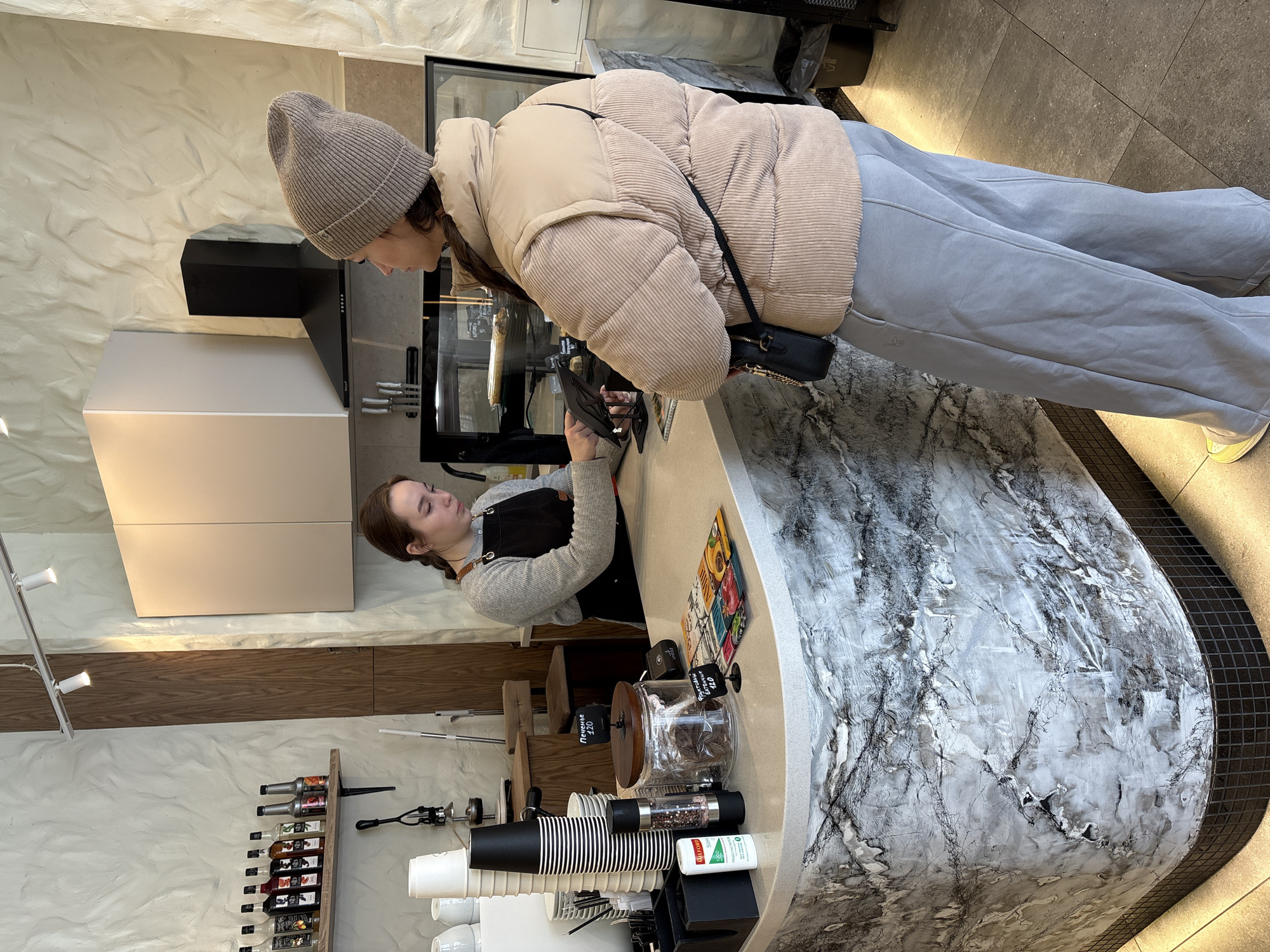 The image shows a person wearing a beige hat, a beige puffer jacket, and gray sweatpants, looking at items in a glass display case in a cafe. A barista stands behind a counter, seemingly waiting to assist the customer.
The cafe has a modern design with a white marble countertop and a variety of coffee-related items such as cups, coffee beans, and syrups on display. The wall behind the barista is white with a textured finish, and there's a wooden cabinet. The location is likely Ulyanovsk, Russia based on metadata.