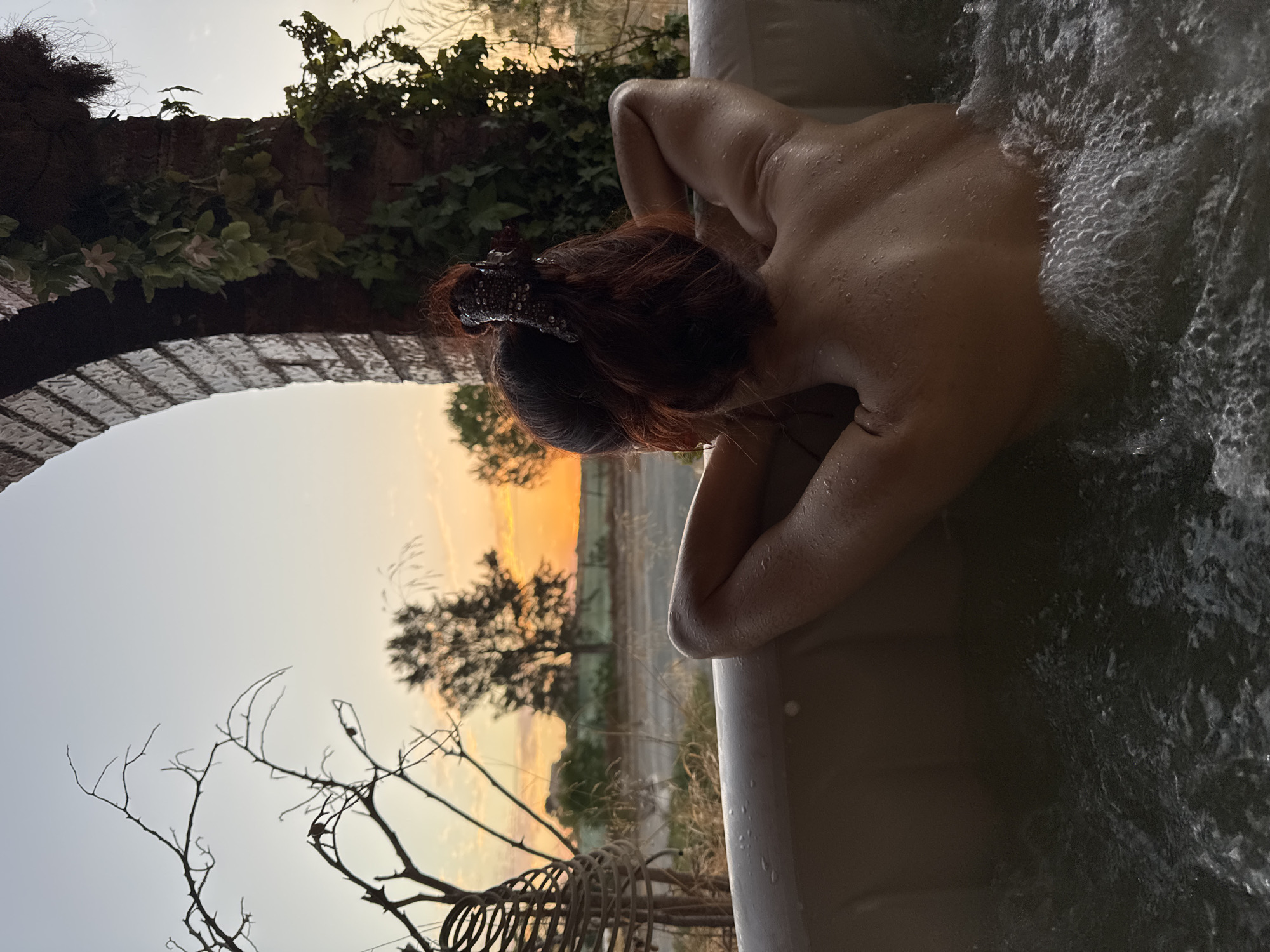 In Corvera, Spain, a person with auburn hair is relaxing in an outdoor hot tub at sunset. The person is facing away from the camera, with their arms behind their head, enjoying the view. The hot tub is filled with bubbling water, and the person's skin is wet, indicating they are submerged in the water. The background features a picturesque landscape with trees, fields, and a vibrant sunset sky. An arched brick structure with hanging greenery is visible to the right of the frame, framing the scene. A wicker decorative object is also present to the left, adding to the rustic ambiance of the location. The overall atmosphere is one of tranquility and relaxation.
