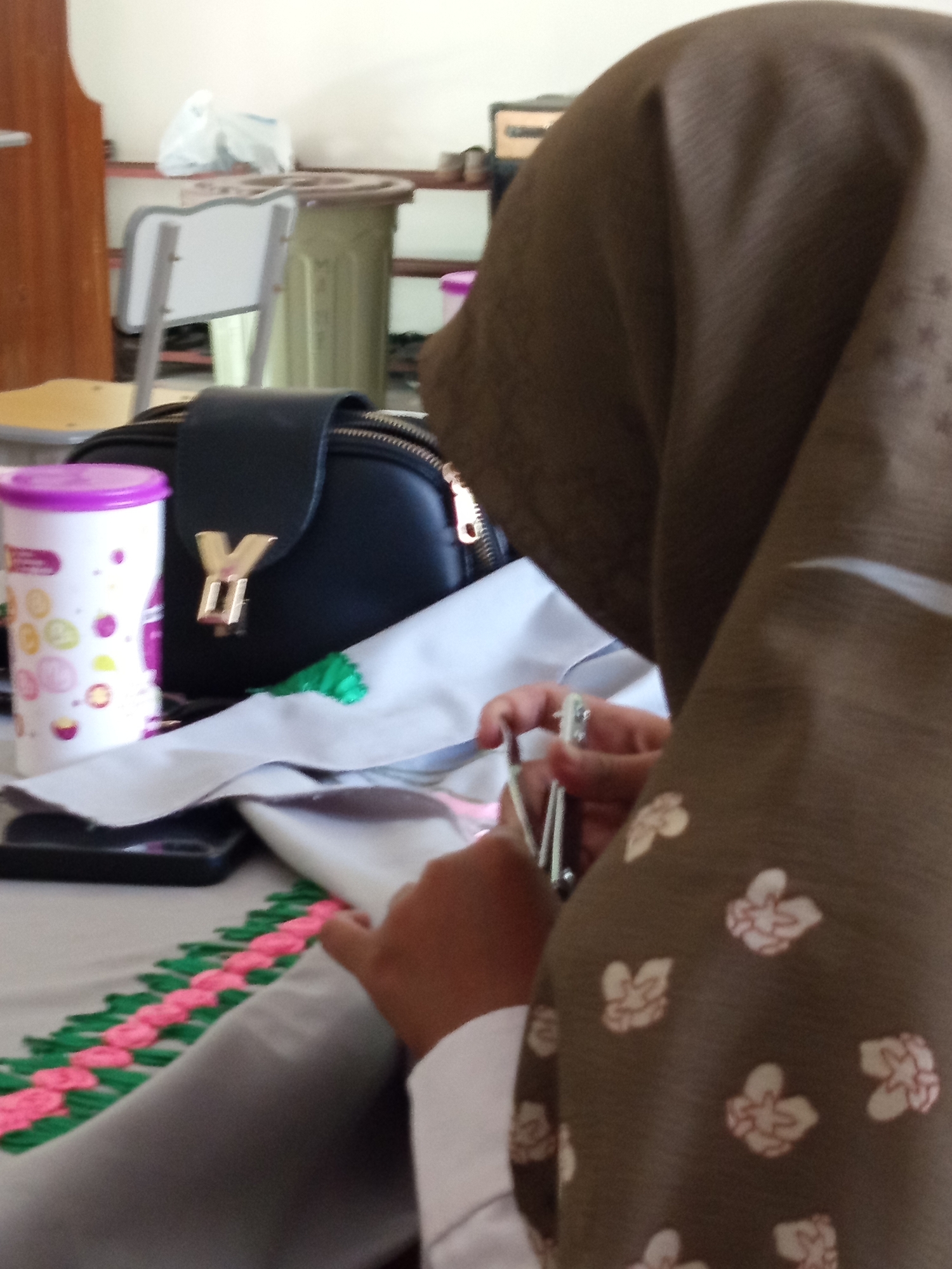 In Lumutan, Indonesia, a close-up shot captures a person wearing a brown hijab with a floral pattern, engaged in embroidery. The person's hands are visible, holding scissors and working on a piece of white fabric adorned with a green and pink braided border. A black purse with a gold accent and a white cup with colorful patterns are placed nearby on a table. The background is blurred, showing hints of a classroom or office setting with wooden furniture and white walls.