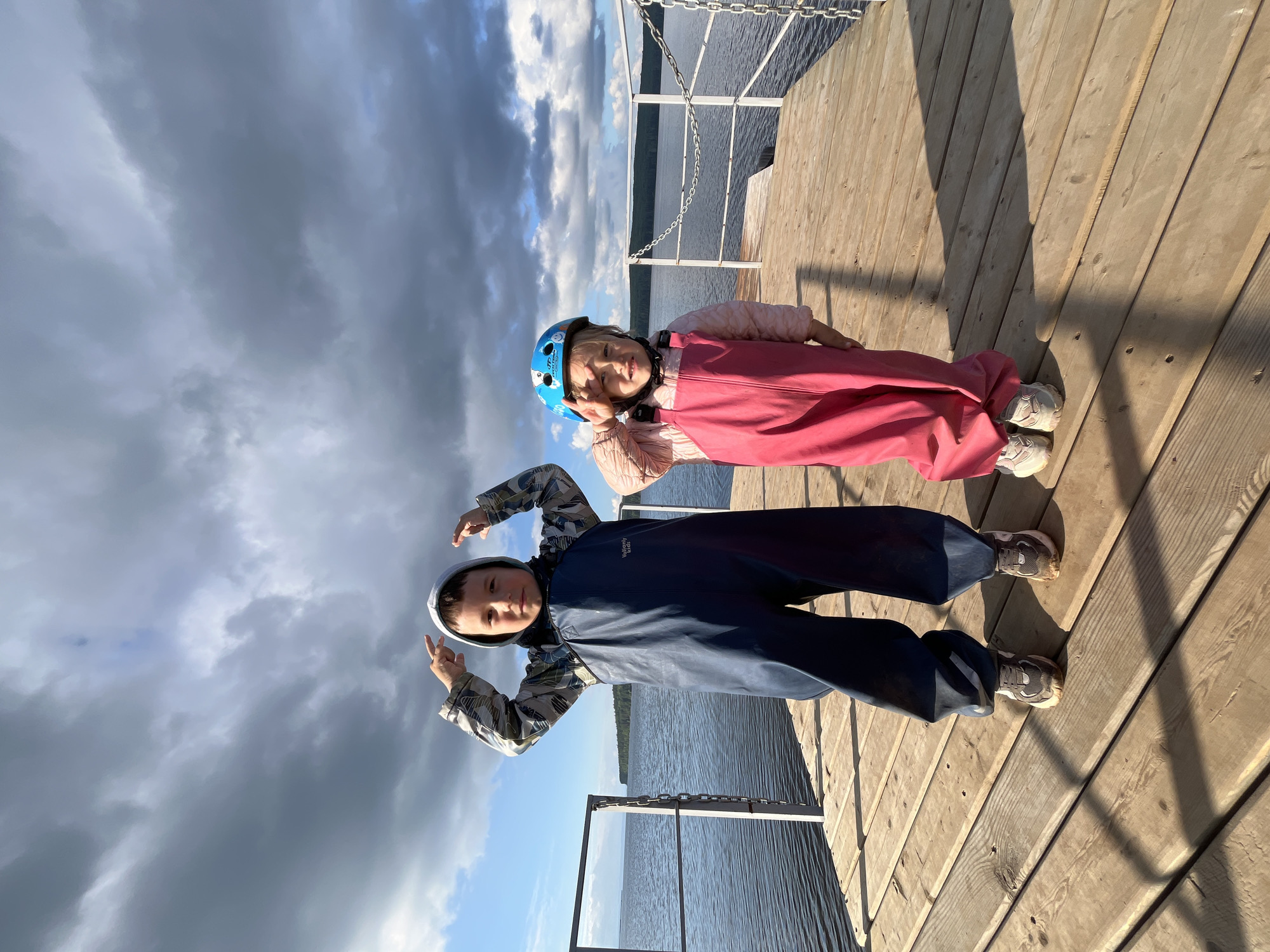 Two young children stand on a wooden pier by a body of water. The child on the left, a boy, wears a grey and green patterned hooded jacket over dark blue overalls and grey and white sneakers. He has his hands raised to his head, fingers pointing upwards. The child on the right, a girl, wears a light pink jacket over bright pink overalls and light-colored sneakers. She has a blue helmet on her head and her right hand is raised to her forehead, with two fingers extended. The pier is made of light-colored wooden planks. In the background, the water is calm and reflects the overcast sky, which is filled with dark grey clouds. A white metal railing with a chain runs along the edge of the pier. The scene appears to be set in a natural outdoor environment, likely a lake or river, with a distant shoreline visible. The overall lighting suggests it is daytime, possibly late afternoon, with diffused sunlight. The location is identified as the city of Chaikovsky, Russia.