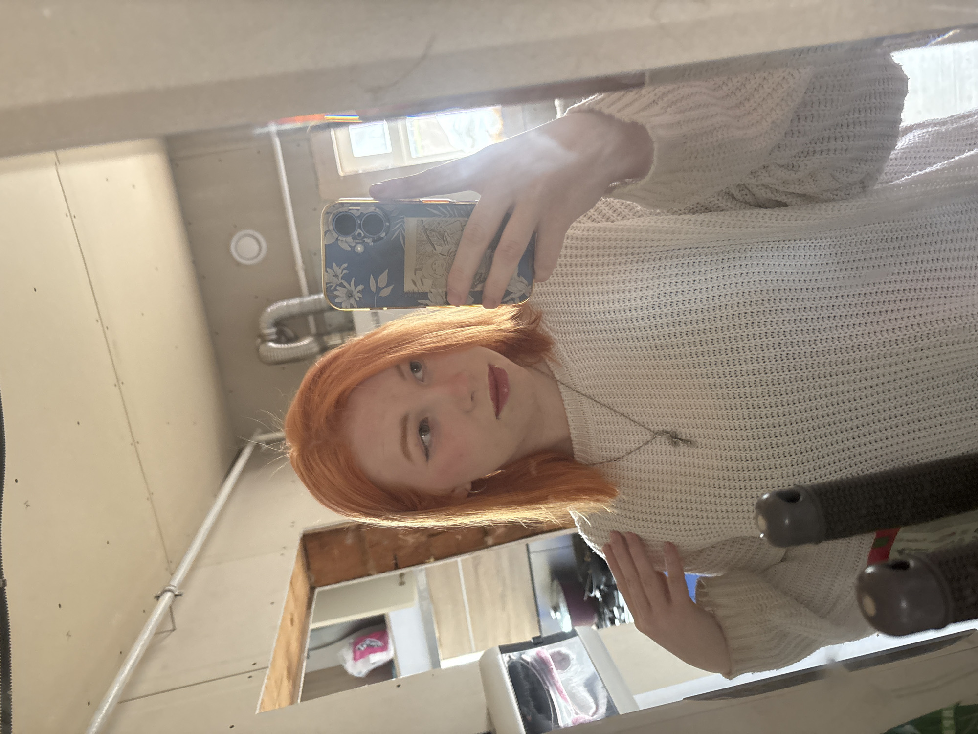 A mirror selfie captures a young person with bright orange-red, shoulder-length hair and fair skin. They are wearing a light-colored, possibly cream or off-white, knitted sweater with a textured pattern and a thin silver necklace with a small pendant. The person holds a smartphone with a dark blue case, adorned with a white floral or leaf pattern, to capture their reflection. The phone case features two prominent camera lenses on the back.
The person has a neutral to slightly contemplative expression, with a subtle hint of a smile, and is wearing lipstick. Their gaze is directed towards the phone's lens.
The background, visible as a reflection in the mirror, suggests an unfinished or utility-style interior. The walls appear to be bare drywall or unpainted wood panels, with visible seams and fasteners. A bright window or light source is reflected in the upper background, illuminating the scene. Other elements reflected in the background include a white circular vent or light fixture on a wall, and silver-colored pipes (both corrugated and straight) attached to the walls and ceiling. Below the person's reflection, there are hints of shelves or storage units containing items, including a pink-wrapped package and other dark objects. On the far right, two dark, textured cylindrical objects, possibly handles, are partially visible. The bright lighting indicates it is daytime.