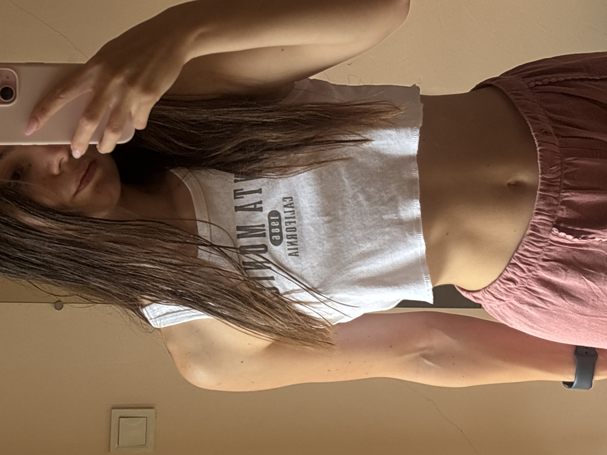 A young woman with long, dark, wet hair is taking a mirror selfie in what appears to be a residential interior. She is wearing a white, cropped tank top with black lettering and pink, high-waisted, loose-fitting pants. Her left arm is visible, and she is wearing a dark smartwatch. The background is a plain, peachy-toned wall with a white light switch on the left side of the frame. The woman is holding a pink smartphone in her right hand, angled to capture the selfie. The lighting suggests it is daytime. The image is likely taken in Varna, Bulgaria, given the context provided.