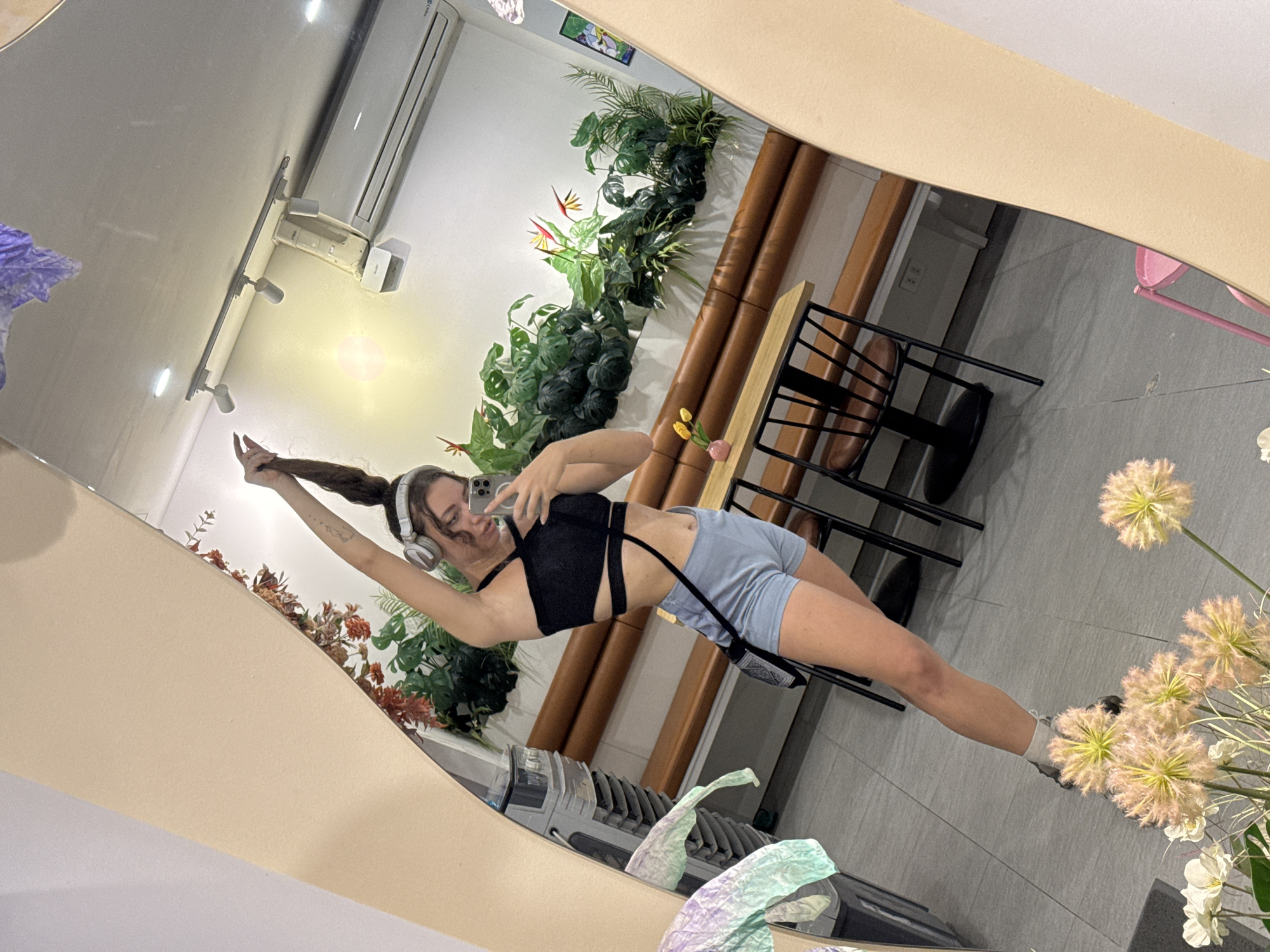 A young woman is taking a mirror selfie in a cafe. She is wearing headphones, a black crop top, and light blue shorts. Her hair is pulled up in a high ponytail, and she is holding her phone in her right hand. The cafe appears to be modern and has a lot of plants. In the reflection of the mirror, we can see a table with two chairs, a bench, and an air conditioner unit. There are also some flowers in the foreground. The cafe is located in Hanoi, Vietnam.
