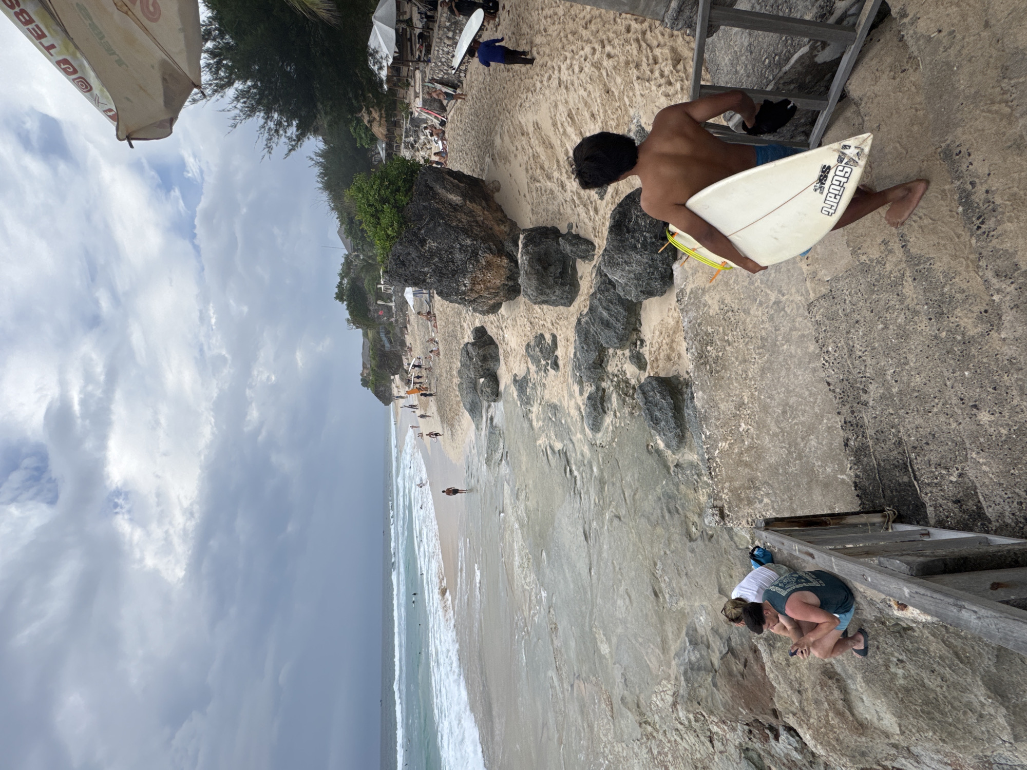 A wide-angle shot captures a busy beach scene in Kuta Selatan, Indonesia, under a cloudy sky. The foreground features a young man with dark hair, shirtless and wearing blue shorts, walking up concrete stairs while carrying a white surfboard. To his left, on rocky terrain, two individuals are seated; one is a male in a teal tank top and blue shorts, looking at his phone, and the other is a female in a white top and grey shorts.
The midground of the image shows a sandy beach dotted with rocks, with several people scattered across it, some walking and others sitting. A line of surf breaks in the turquoise ocean, with a few surfers visible in the water and some surfboards leaning against structures on the beach. Beach umbrellas and thatched-roof huts are present, suggesting a resort or cafe area. The background consists of the ocean meeting the horizon under a grey, overcast sky with patches of blue peeking through. The overall scene depicts a lively beach environment with people enjoying various activities.