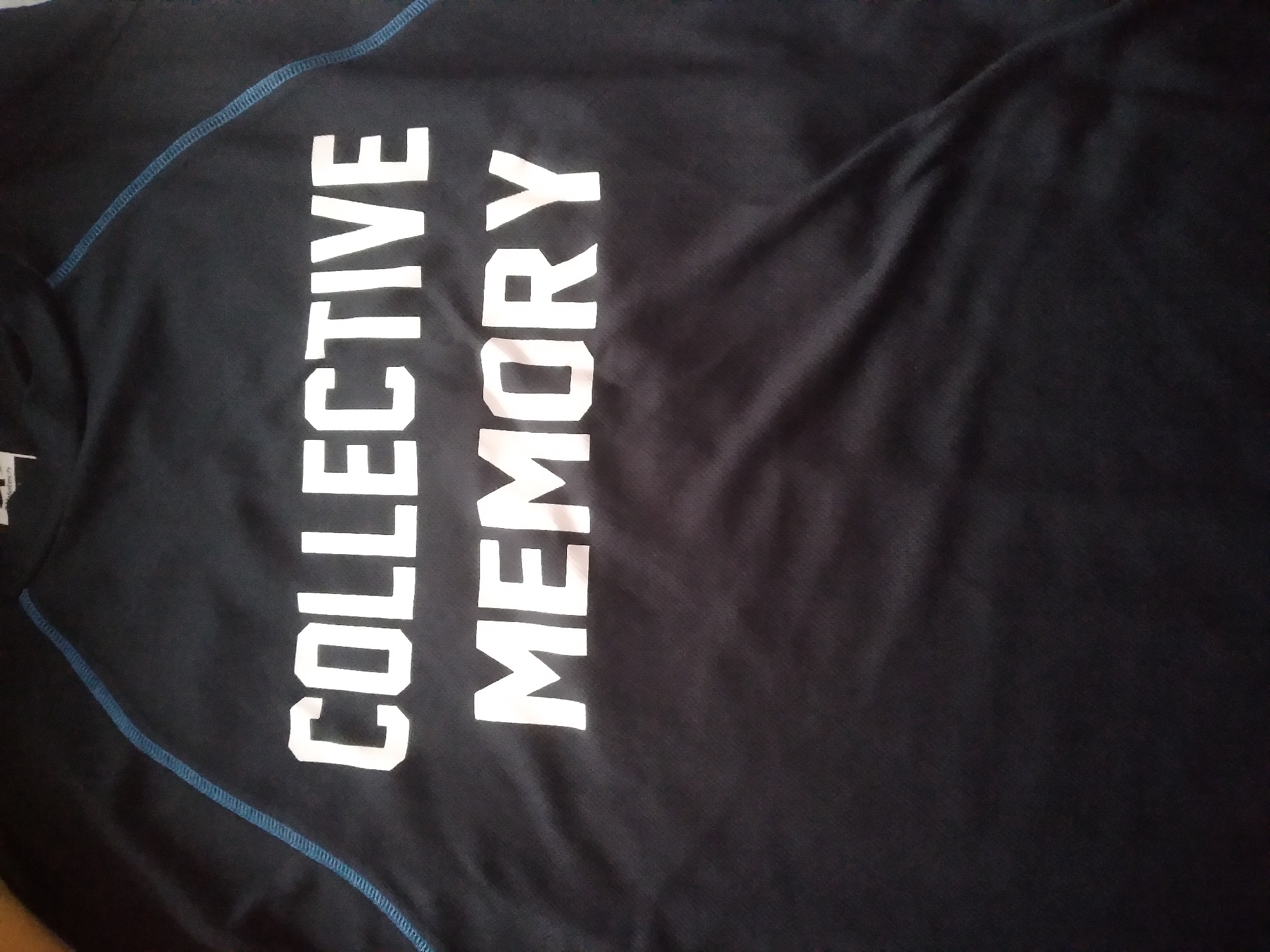 The image shows a close-up view of a black t-shirt with white text. The text reads "COLLECTIVE MEMORY" in two lines. The t-shirt has blue stitching along the seams, visible on the left and right sides of the frame. The fabric appears to be synthetic and slightly wrinkled. There is a small tag at the top center of the shirt, partially visible. The background is entirely black, indicating the t-shirt is laid flat or hung against a dark surface. There are no people, city elements, or indications of a location in Nyeri, Kenya present in the image.