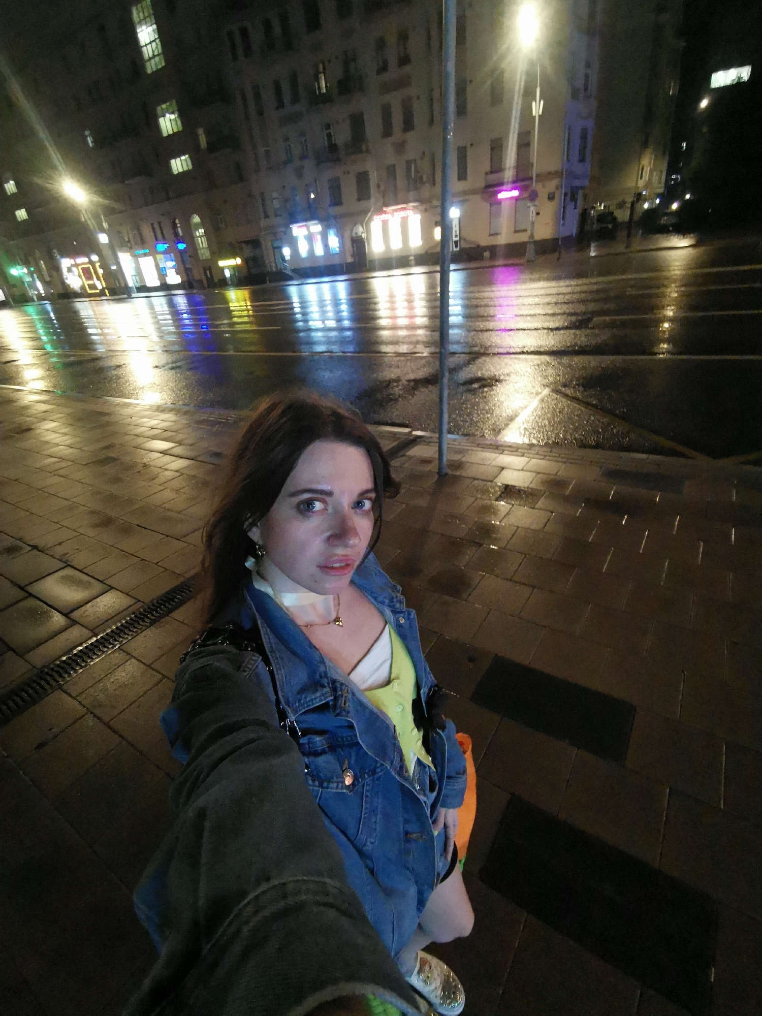 A medium shot shows a woman taking a selfie on a wet city street at night in Unknown, Unknown. The woman is fair-skinned with brown hair. She wears a denim jacket over a light-colored top. Behind her, the street reflects the lights of buildings, with multiple street lights and lit buildings on the other side of the street. The road is wet, reflecting the lights from the buildings. The sidewalk is paved with rectangular bricks, and a metal pole stands near the edge of the sidewalk.