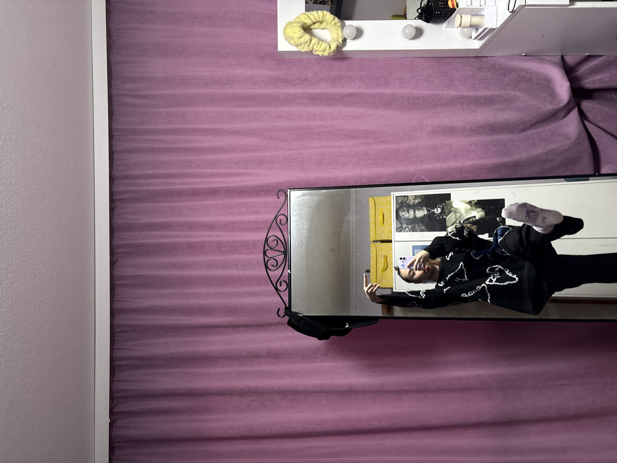 A mirrored reflection of a person wearing a black sweater and pants, posing with their phone, is visible. The reflection is set against a backdrop of a light purple curtain. Above the mirror, a white vanity with lights and a yellow scrunchie can be seen. The location is Прикамский, Russia.