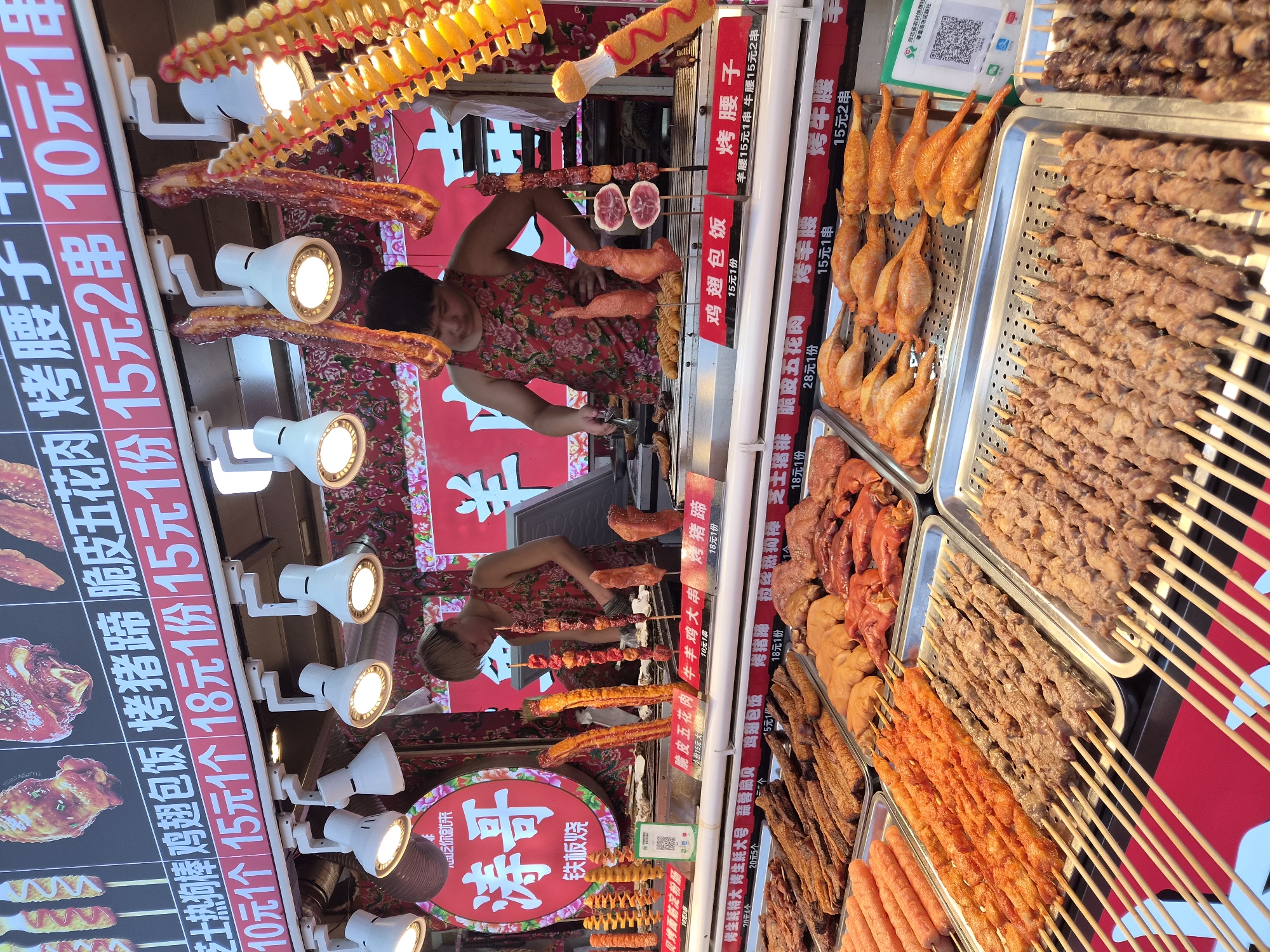 The image depicts an outdoor food stall in Qinhuangdao, China, specializing in grilled skewers and other street food items. Two young individuals, likely vendors, are visible behind the counter. One is actively preparing food on a grill, while the other appears to be attending to the display. The stall is brightly lit by overhead lamps, illuminating a variety of raw and cooked meats, including chicken wings, various cuts of beef and pork, and what appear to be sausages or other processed meats arranged in metal trays. Above the food display, a menu board features Chinese characters and prices, indicating a range of offerings such as "烤猪蹄" (roasted pig trotters), "脆皮五花肉" (crispy pork belly), and "鸡翅包饭" (chicken wings with rice). Hanging from the ceiling are decorative items and more food samples. The overall atmosphere suggests a busy street food environment, common in Chinese cities. The presence of a QR code on a green sign suggests a digital payment option or information portal for the stall.