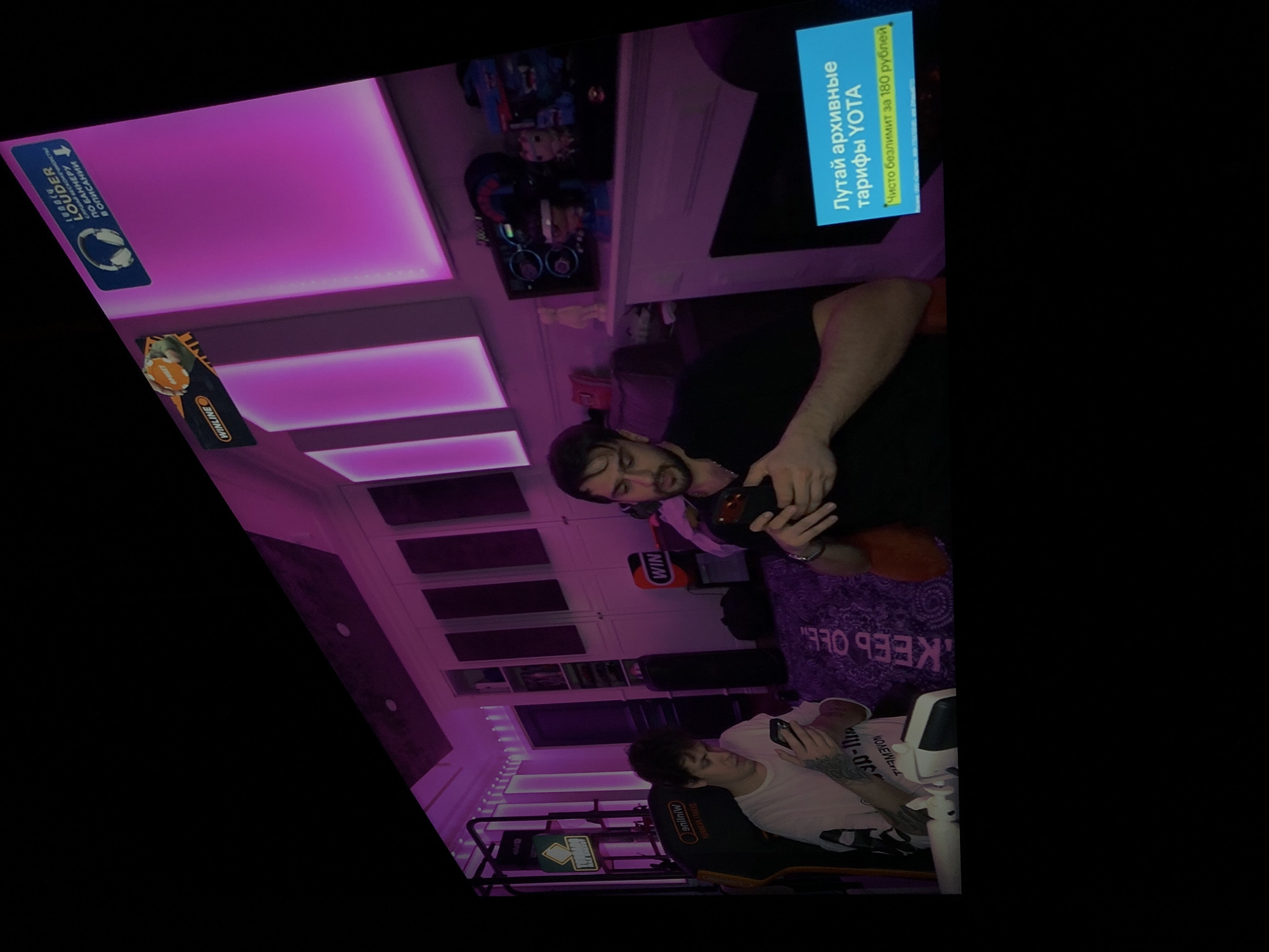 Here is a forensic description of the image:
**Scene:** Interior shot of a room with purple lighting, likely a gaming or streaming setup.
**People:**
* Two men are present. One is seated in a gaming chair using a mobile phone. The other man stands behind him, holding a black bottle.
**Objects:**
* A gaming chair with a "Winline" logo and lettering "КОННЕЛЯ ГУМЭД"
* Mobile phones.
* A black bottle.
* A patterned purple blanket with lettering "KEEP OFF" partially visible.
* Shelves and cabinets line the wall, illuminated by purple lighting.
* A display screen with ad text "Лутай архивные тарифы YOTA. Чисто безлимит за 180 рублей."
* Decorative wall panels.
**Actions:**
* The seated man is using his mobile phone.
* The standing man appears to be holding and examining the bottle.
**Environment:**
* The room is decorated with purple lighting.
* The overall environment suggests a casual, possibly gaming-related space.
There is insufficient information to determine an exact location or city.