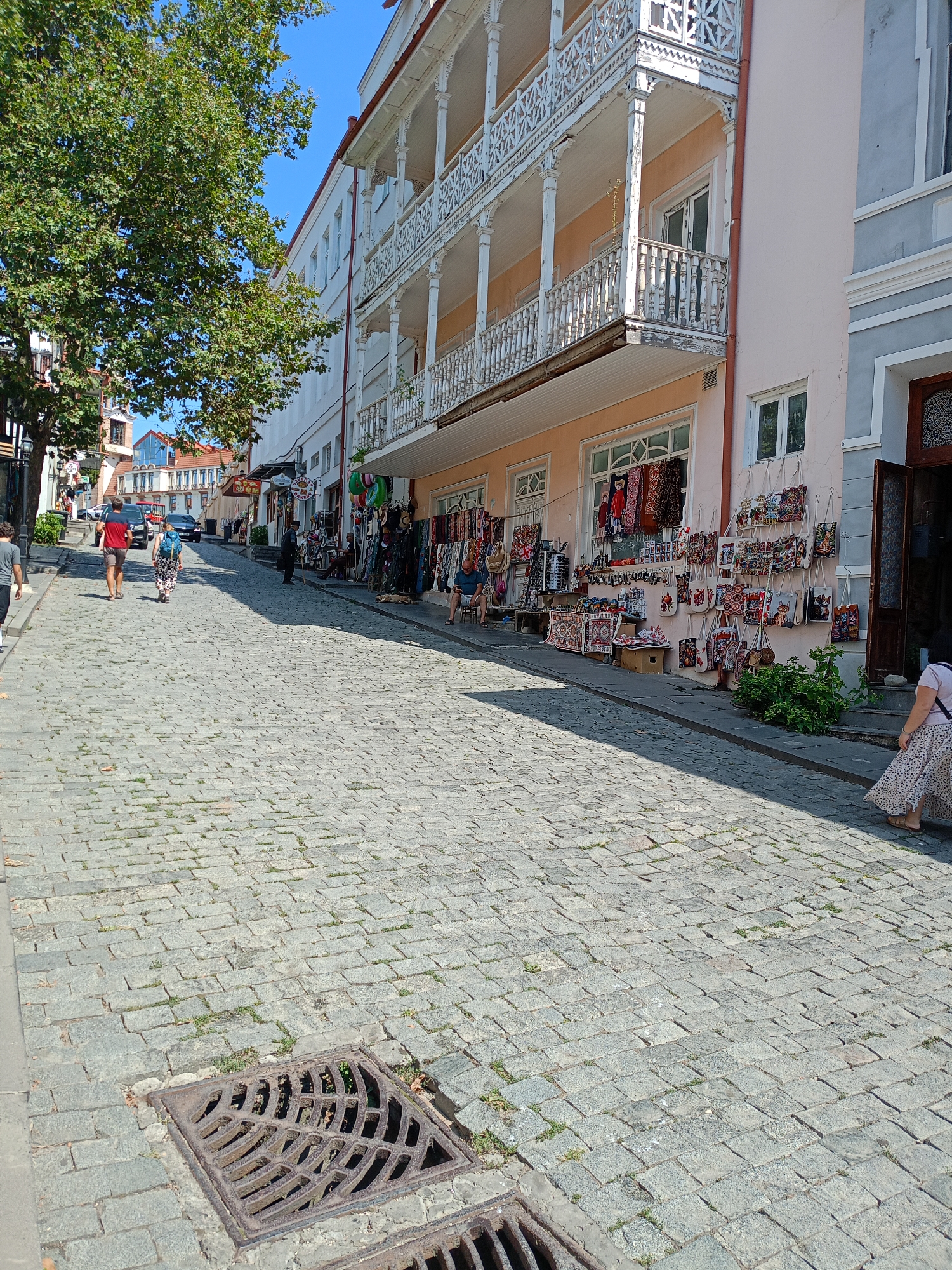 The image depicts a cobblestone street in the city of Sighnaghi, Georgia. The street is lined with buildings featuring traditional Georgian architecture, characterized by ornate wooden balconies and pastel-colored facades. One building on the right displays a shop with various handmade bags and textiles for sale, hanging from its exterior walls. People are seen walking along the street, some browsing the merchandise. A large tree with green foliage shades a portion of the left side of the street, while the sky above is clear and blue. The scene captures a vibrant street life with a blend of historical architecture and local commerce.