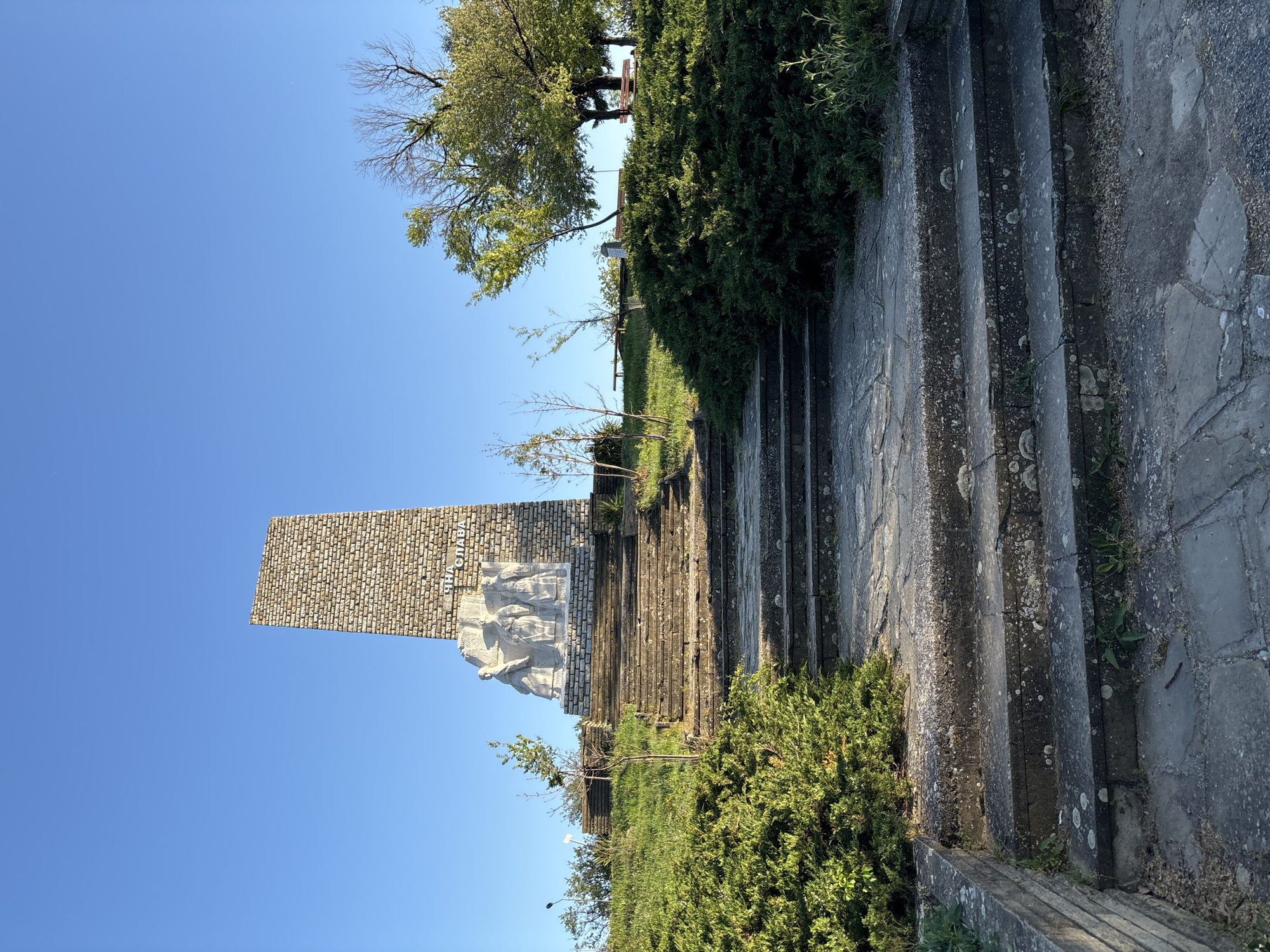 The image depicts a monument situated on a hill or elevated area. The monument consists of a tall, brick-like structure topped with a sculpture of figures. Inscribed on the monument in Cyrillic script is "В ИМЕТО НА СЛАВАТА". A set of stone stairs leads up to the monument, lined with vegetation, including trees and bushes. The sky is a clear blue. The city is Unknown, Unknown.