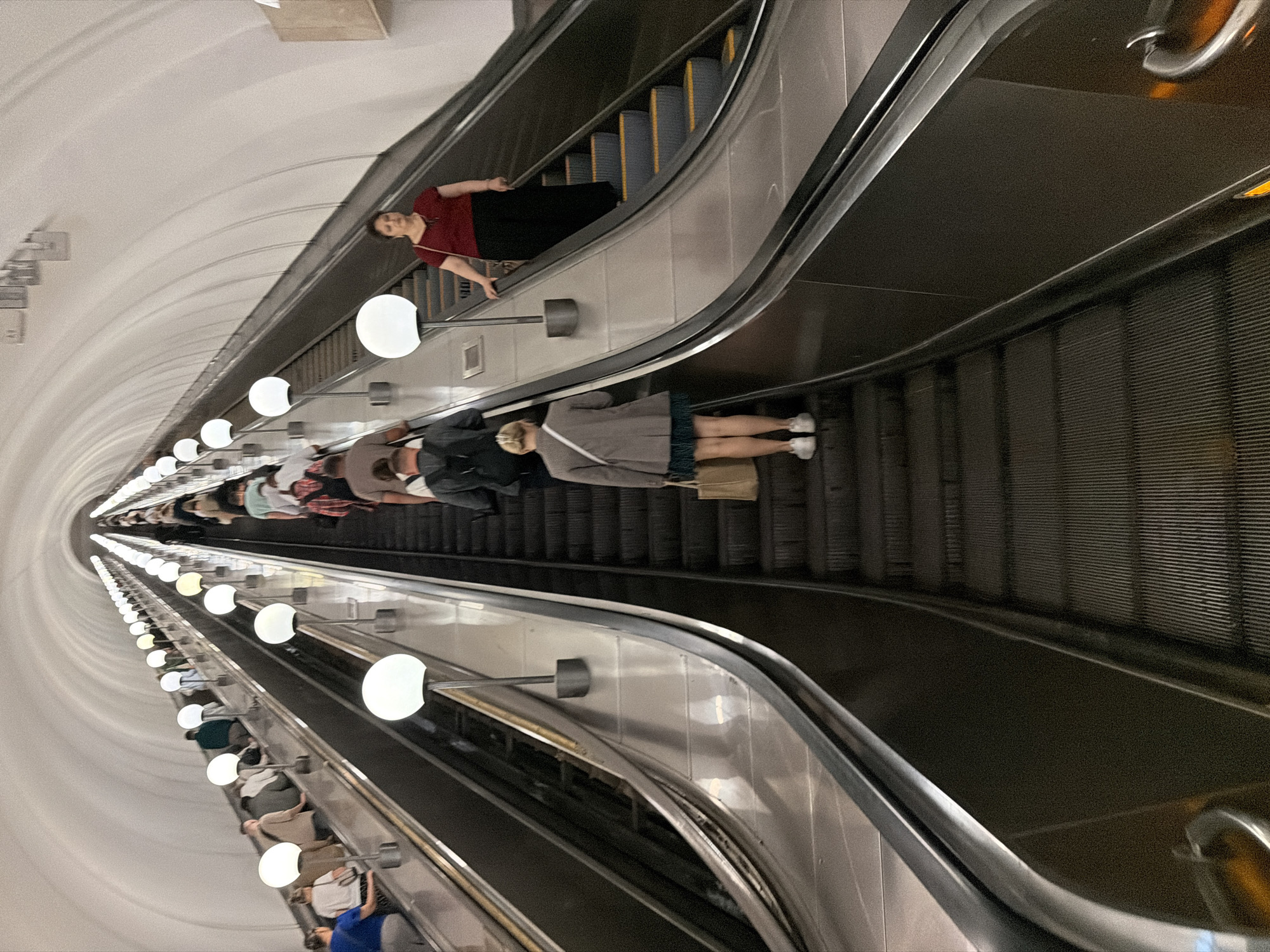 An underground escalator in an unknown city station features two parallel sets of escalators ascending into a cavernous, brightly lit tunnel. Multiple individuals are seen on both escalators, some ascending and others descending. On the right escalator, a woman in a red shirt and black pants is descending, while on the left escalator, a woman in a grey jacket and short skirt is ascending, carrying a beige tote bag. The station architecture is characterized by a curved, white ceiling and numerous large, spherical white lights illuminating the scene. The escalators themselves are made of metal with ribbed treads, and handrails are visible.