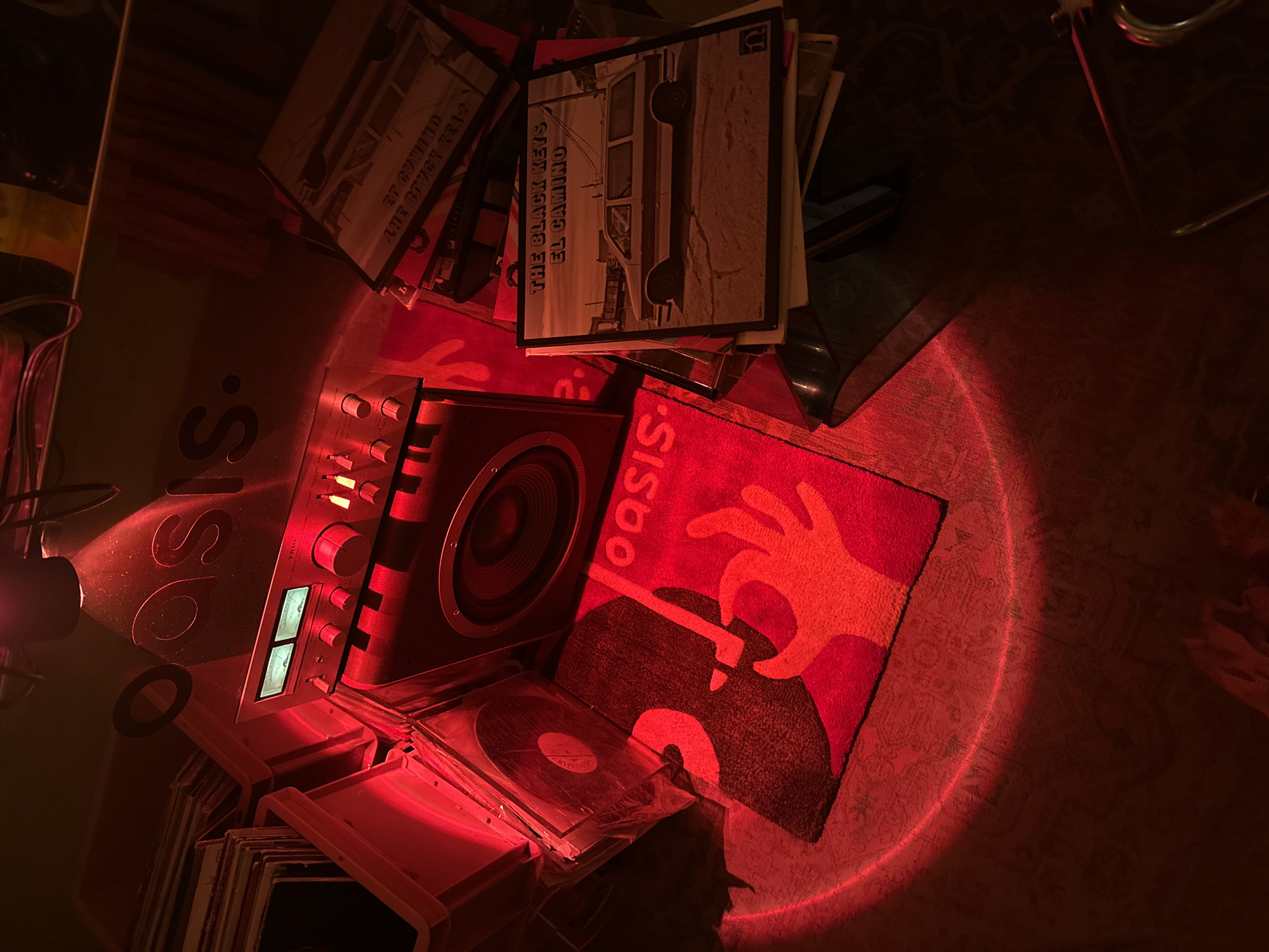 The image shows a collection of vinyl records and audio equipment illuminated by a red light. Visible record covers include "El Camino" by The Black Keys. The environment includes a rug with a hand and record design, and a turntable. The location is unknown.