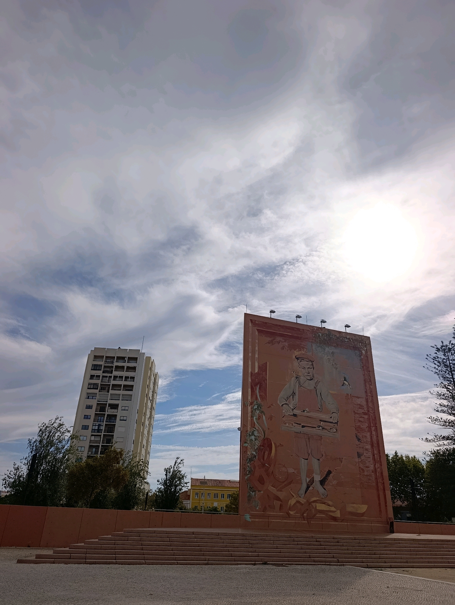 A tall, white residential building stands to the left, its numerous windows and balconies visible. To the right, a large, vertical mural depicts a stylized young boy in overalls, carrying a toolbox. The mural is set against a pale orange background with some abstract shapes. In the foreground, there are tiered steps leading up to the mural. Trees line the middle ground, with a bright yellow building visible in the distance behind the trees. The sky is cloudy, with the sun shining brightly, causing some glare. The overall setting appears to be an urban public space in Setúbal, Portugal.