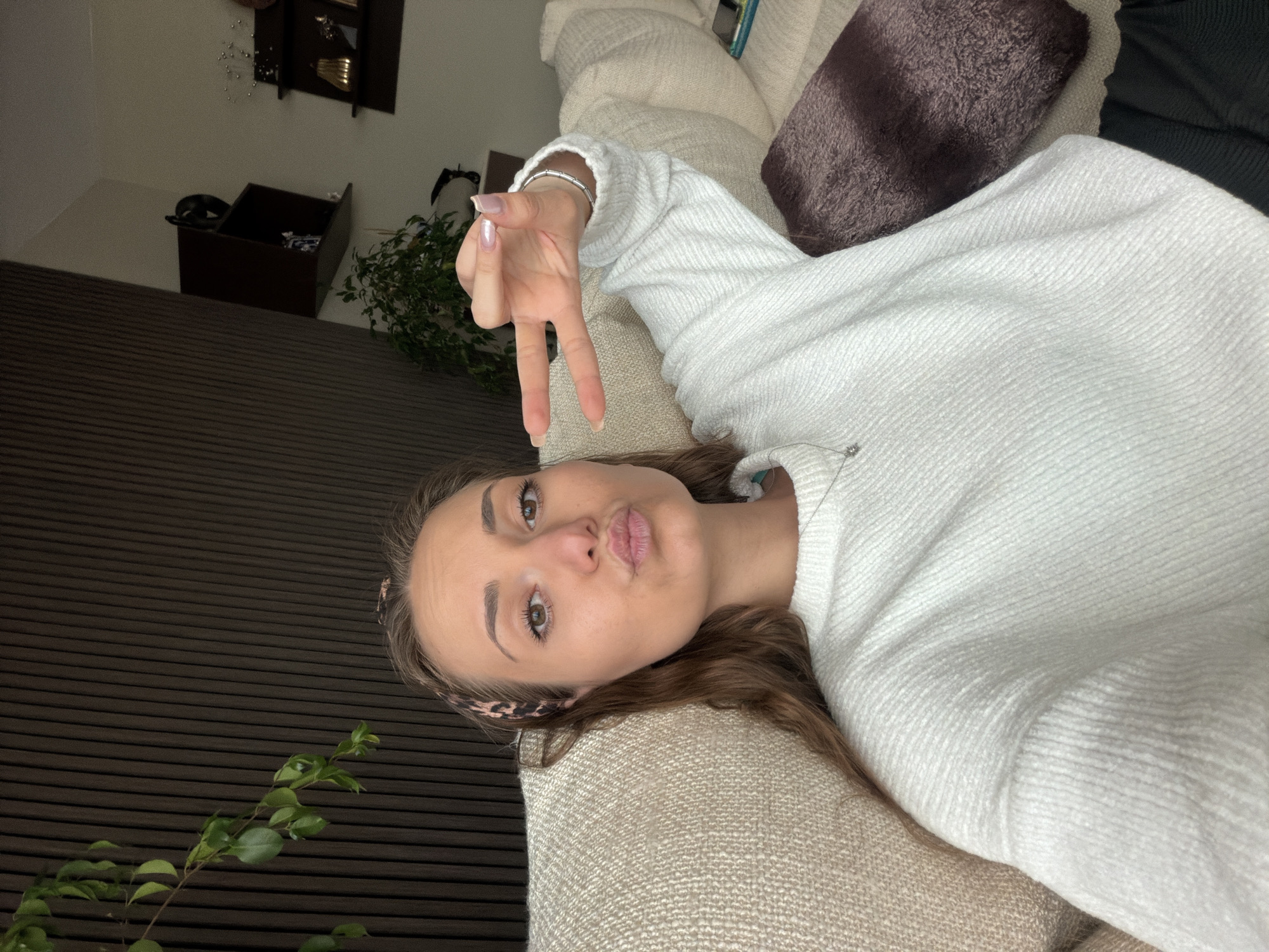 The image shows a young woman lying on a couch in what appears to be an indoor setting. She is wearing a white sweater and a leopard print headband. Her hair is brown. She is making a peace sign with her hand and puckering her lips. A plant is visible in the foreground and background. The location is not possible to identify as being in Buhovo, Bulgaria from the provided information.